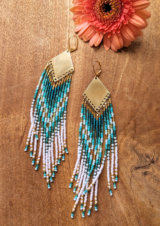 Moon & Milk - Long beaded earrings with white, turquoise, and gold beads in a chevron pattern, gold diamond-shaped tops. Photographed on a wooden surface with a pink flower.