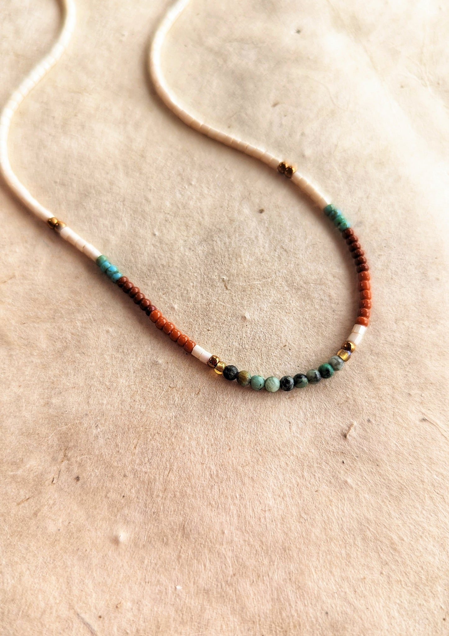 Moon & Milk - Delicate beaded choker necklace with ivory, brown, turquoise, and gold beads, arranged in a simple pattern. Photographed on a beige background.