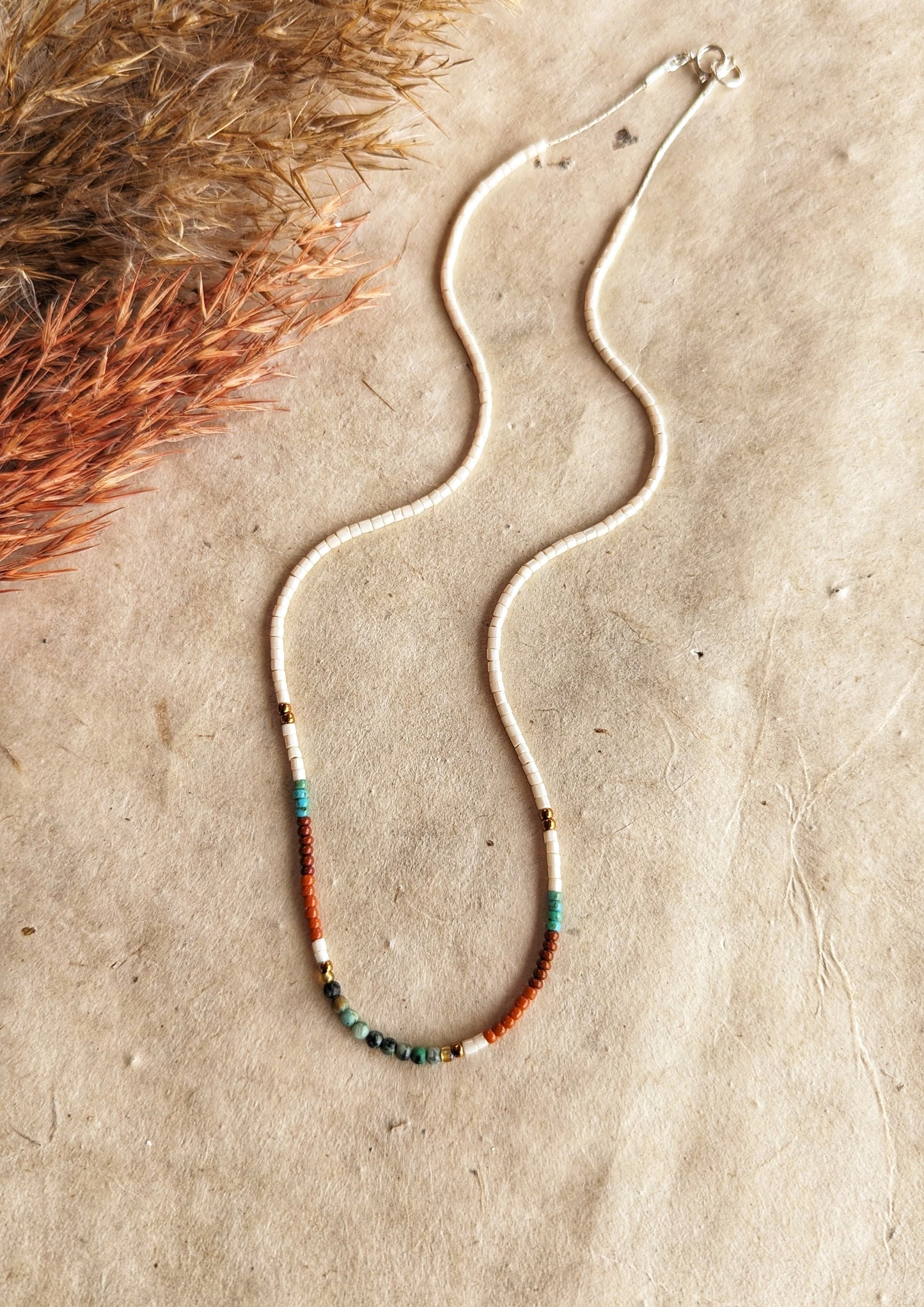Moon & Milk - Delicate beaded choker necklace with ivory, brown, turquoise, and gold beads, arranged in a simple pattern. Photographed on a beige background.