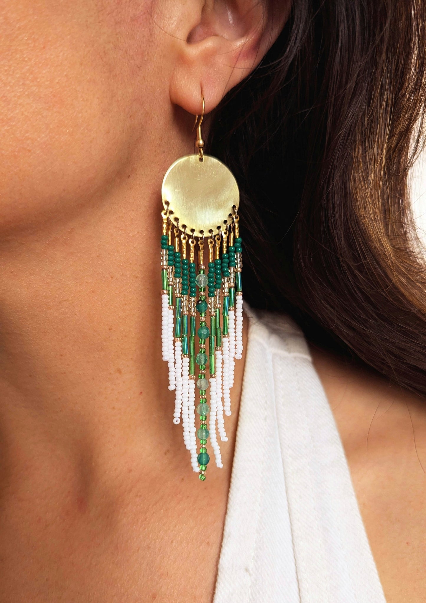 Gold dangling seed bead earring with white and green beaded strands, hanging from a large circular golden brass disc. Worn by a woman with dark hair.