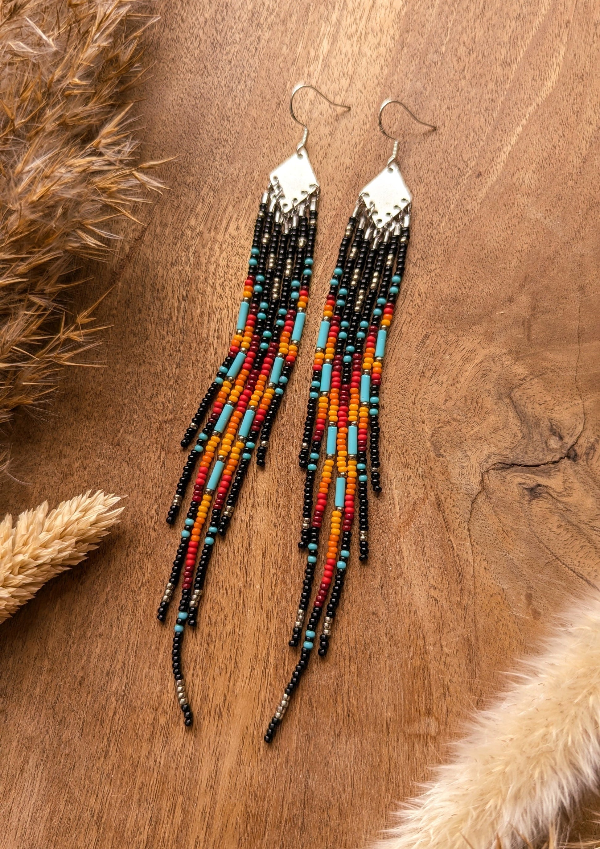 Handcrafted beaded earrings with a vibrant fringe design. The beads feature a bold mix of black, red, yellow, turquoise, and metallic accents, creating a striking, colorful pattern.