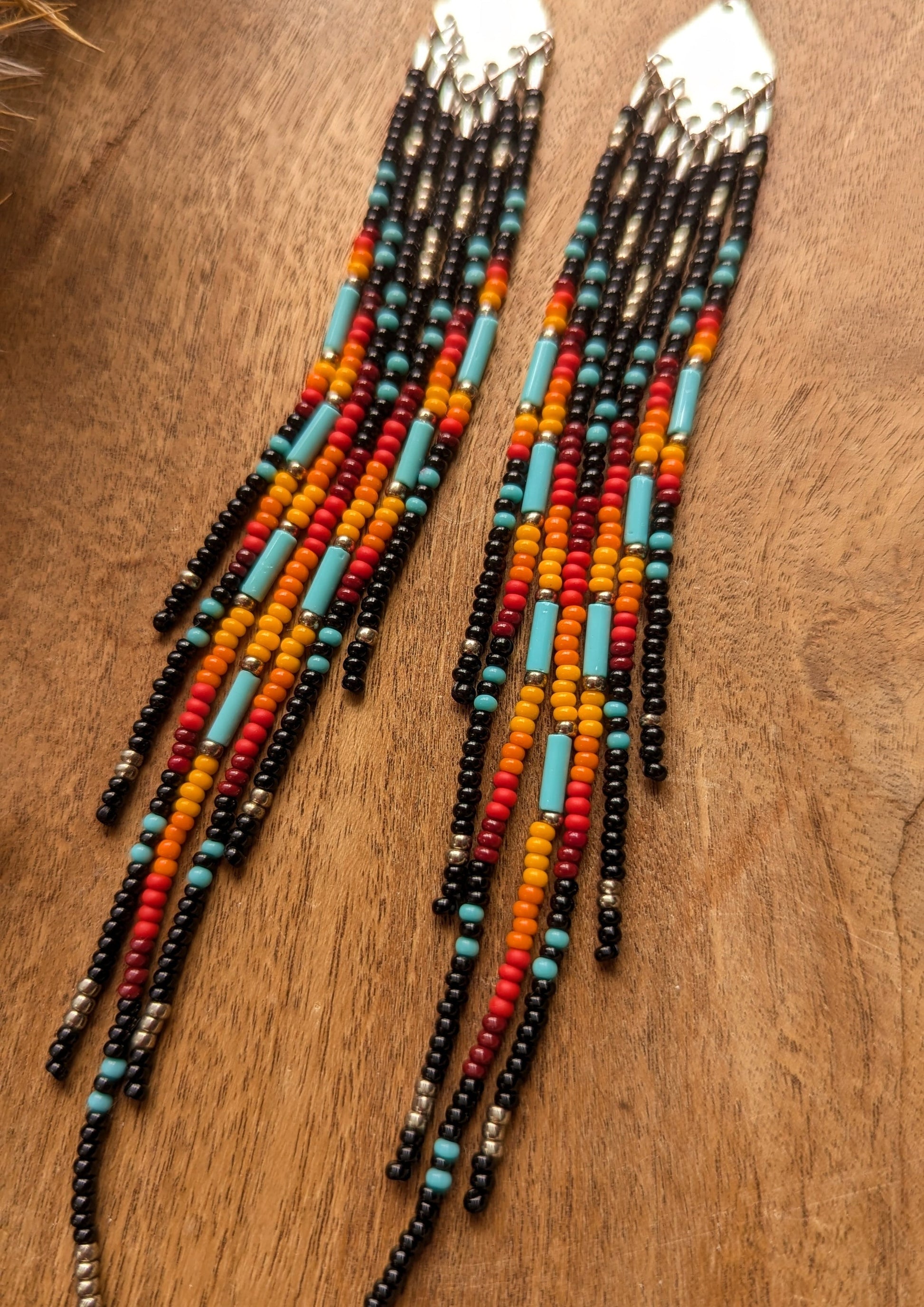 Handcrafted beaded earrings with a vibrant fringe design. The beads feature a bold mix of black, red, yellow, turquoise, and metallic accents, creating a striking, colorful pattern.