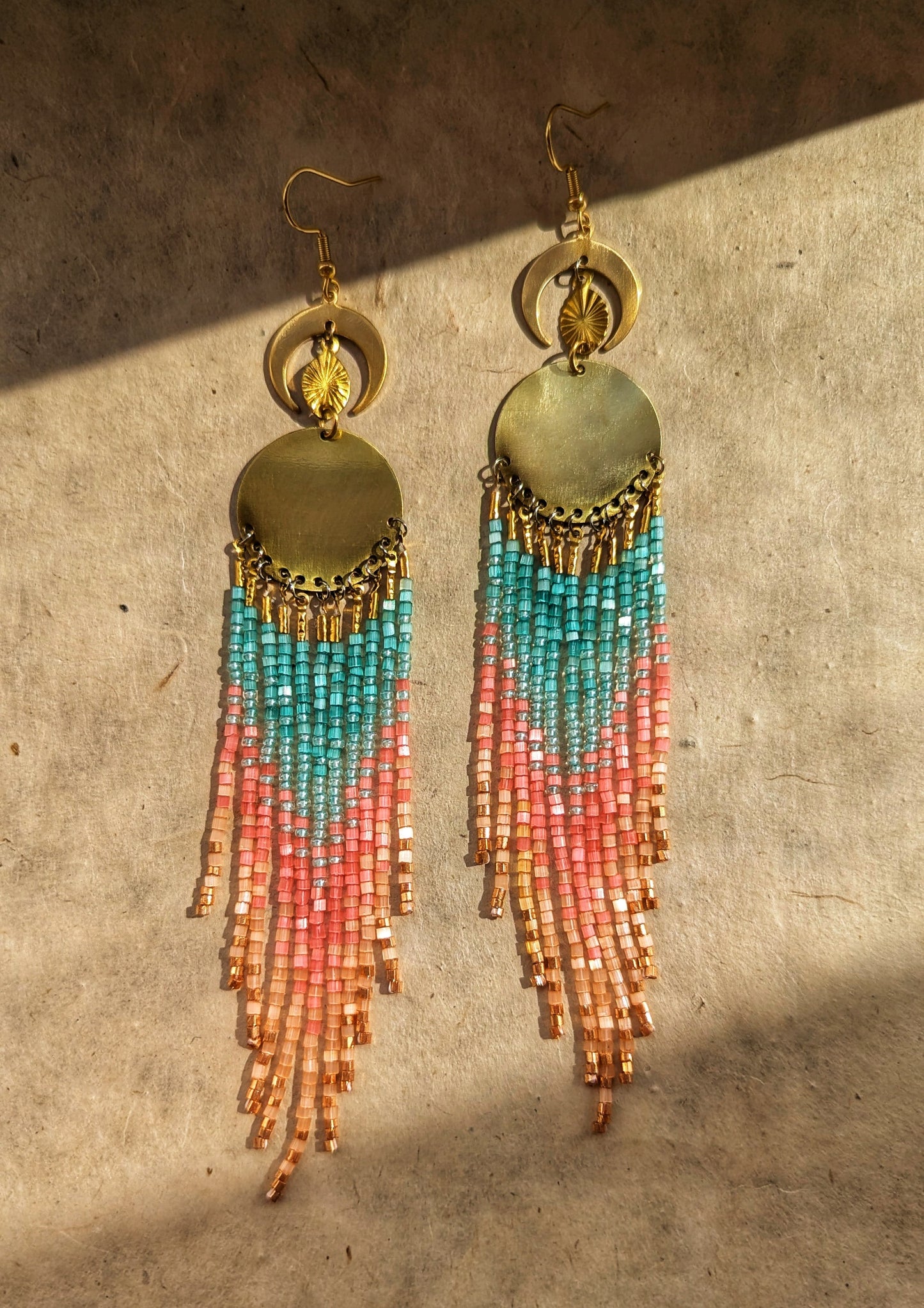 Beaded fringe earrings adorned with crescent and sun details, showcasing long, cascading beaded strands in soft shades of turquoise, pink, and peach, placed gracefully against a textured background.- Moon & Milk
