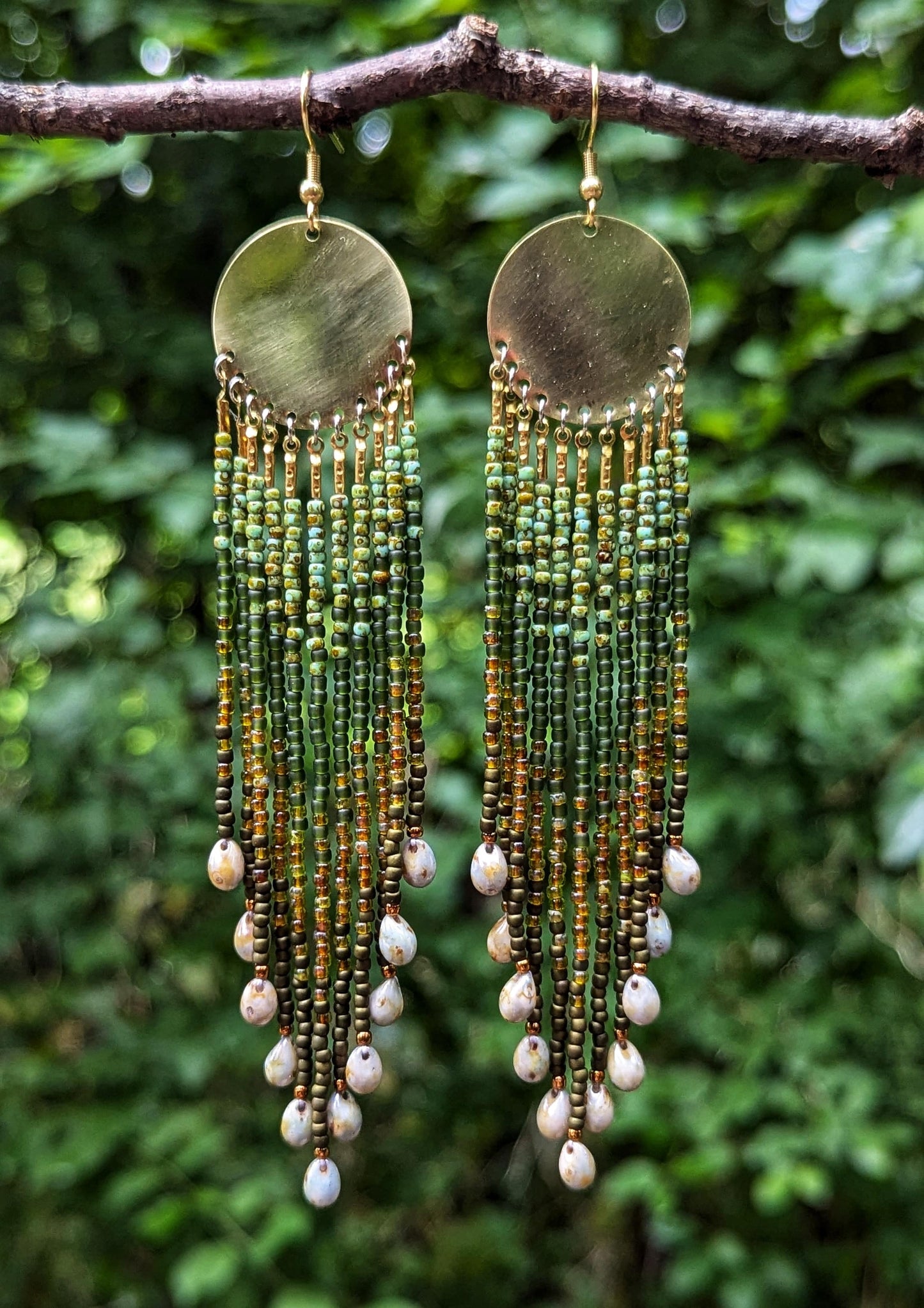 Green and gold seed bead earrings with glass tearsrops and brass disks hanging from a branch in a lush green forest.