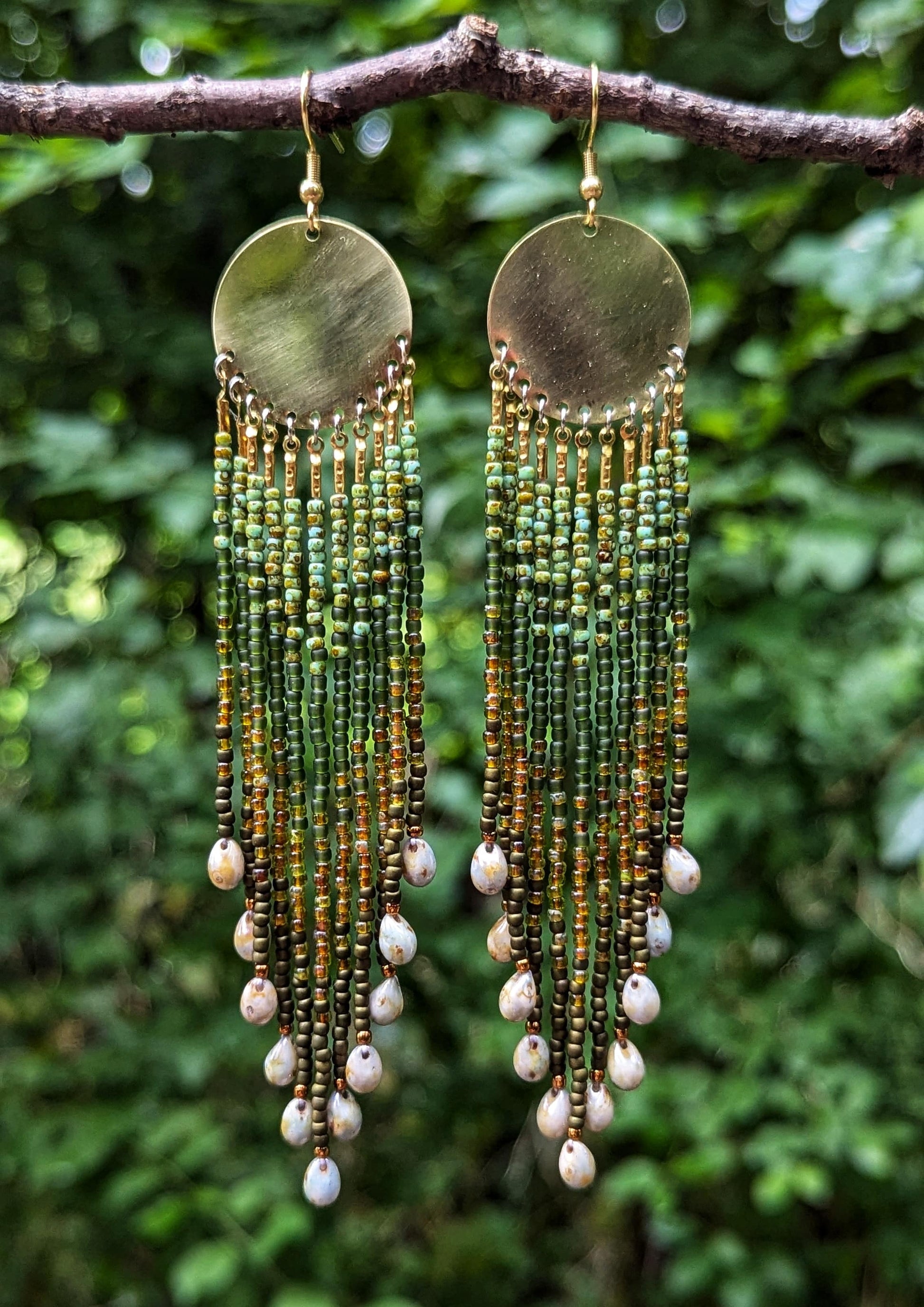 Green and gold seed bead earrings with glass tearsrops and brass disks hanging from a branch in a lush green forest.