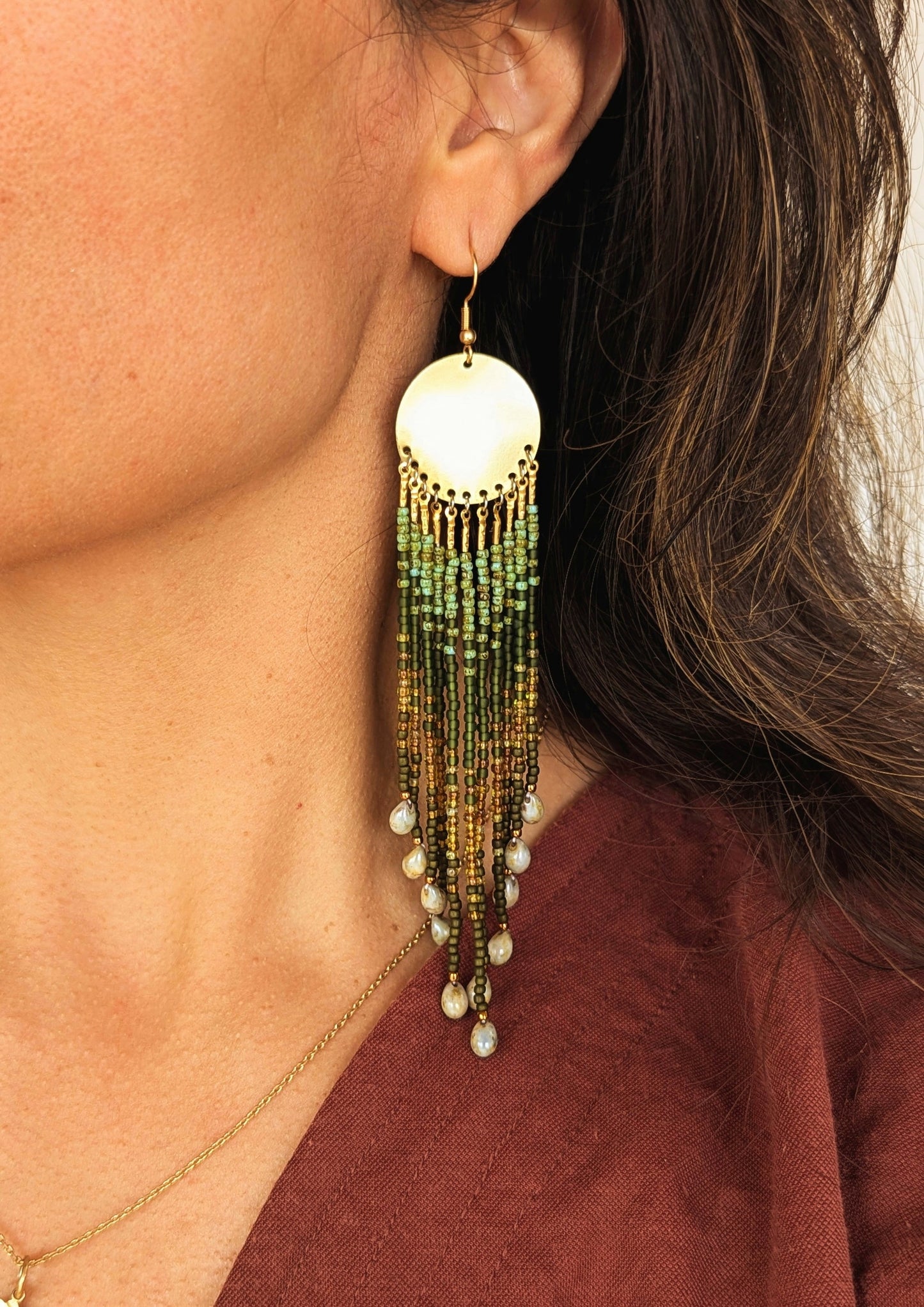 Close-up of a woman wearing long beaded green earrings with brass disks and glass teardrops, styled with a burgundy top.