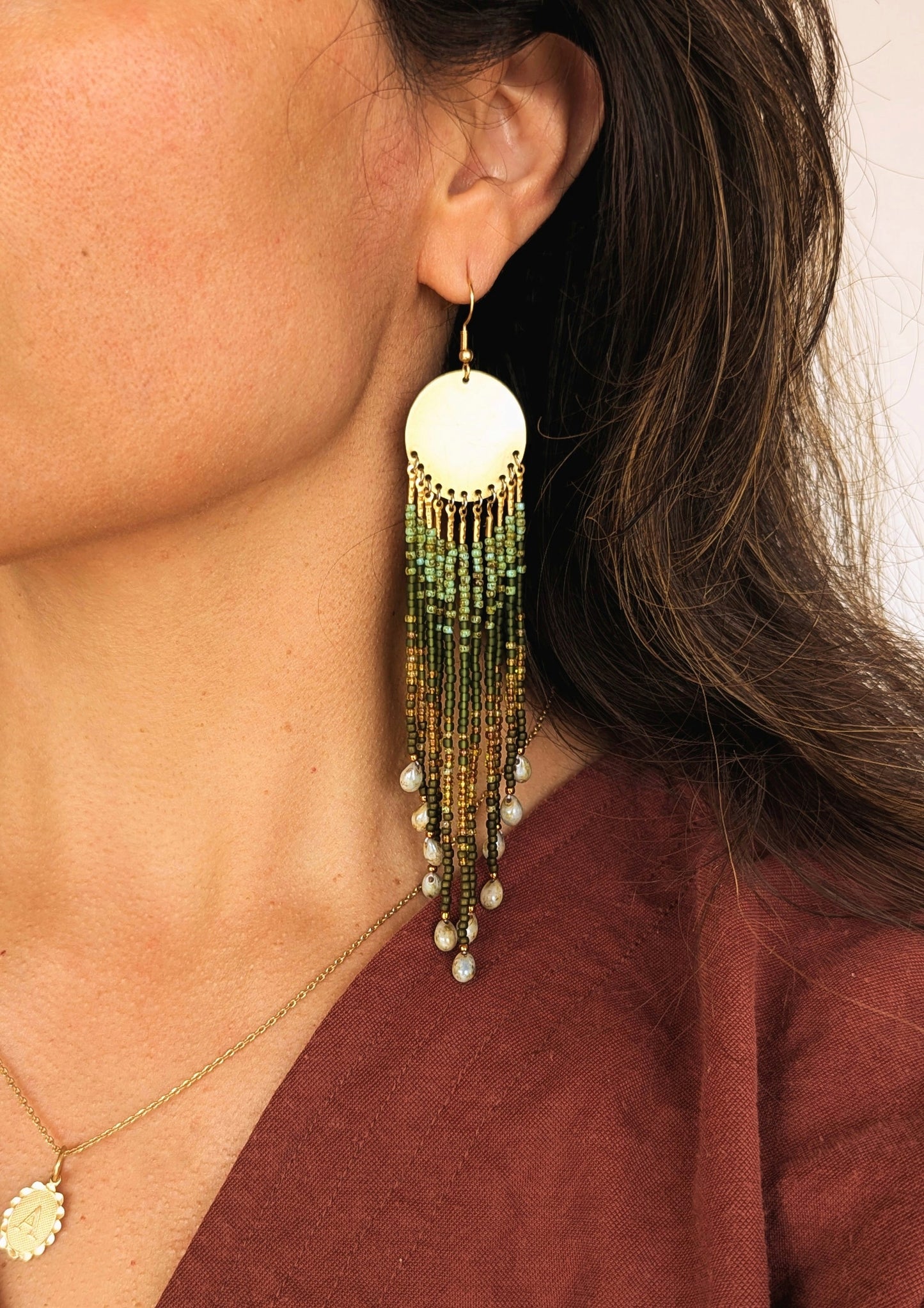 Close-up of a woman wearing long beaded green earrings with brass disks and glass teardrops, styled with a burgundy top.