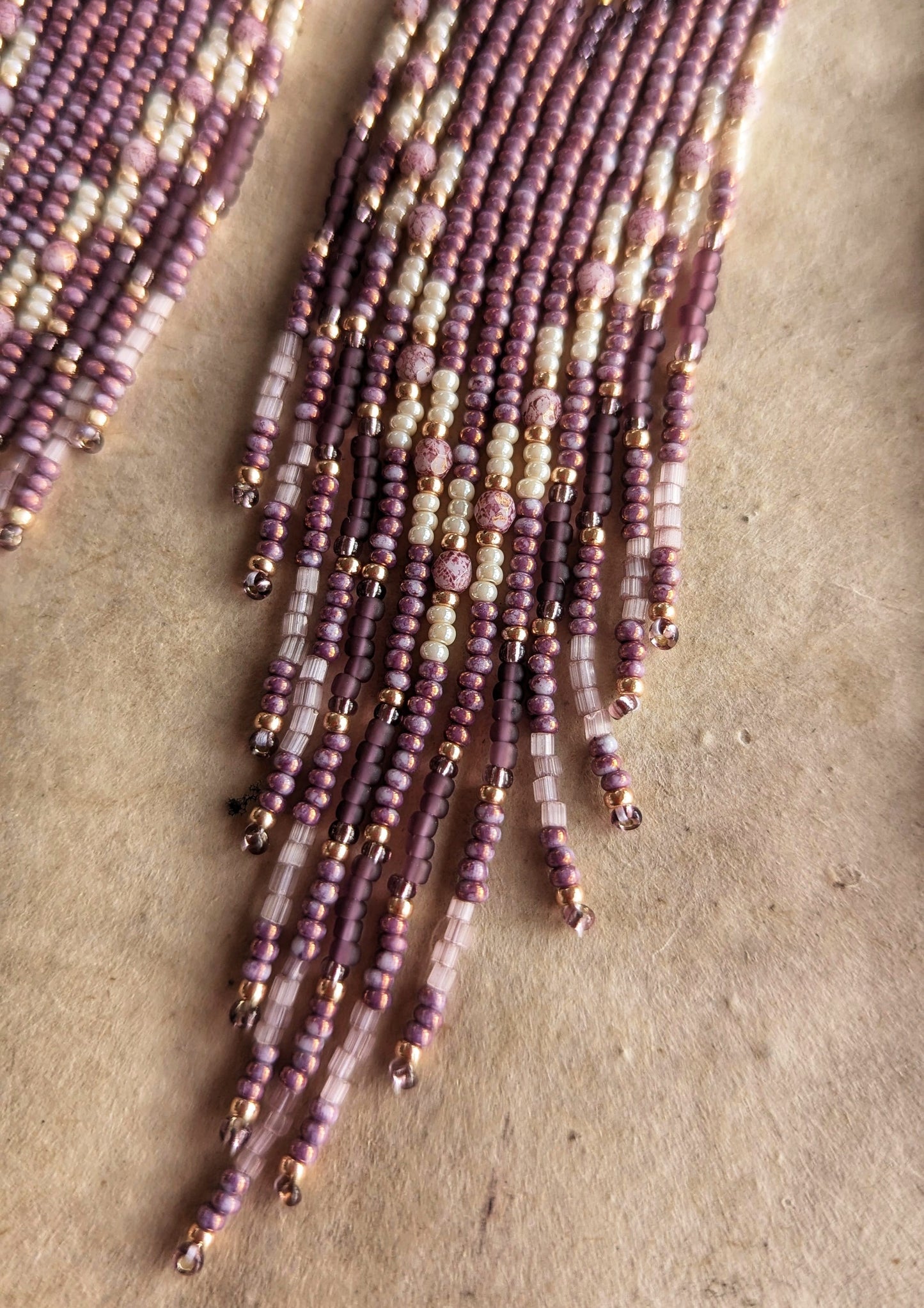 Moon & Milk - Close-up view of intricate beading on earrings featuring delicate strands of purple, cream, and gold beads, artistically laid on a soft, textured background.