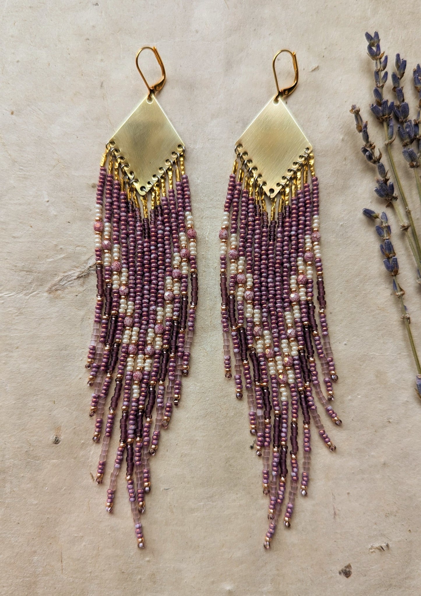 Moon & Milk - beaded earrings featuring purple and cream strands cascading from a gold diamond-shaped brass pendant, displayed on a neutral background with sprigs of lavender.