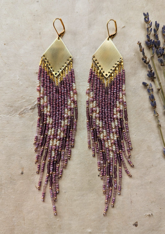 Moon & Milk - beaded earrings featuring purple and cream strands cascading from a gold diamond-shaped brass pendant, displayed on a neutral background with sprigs of lavender.