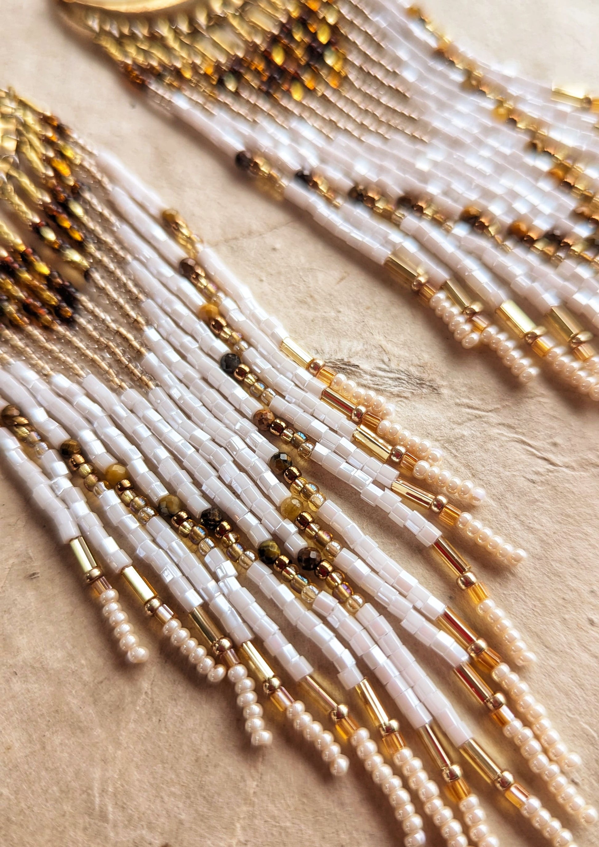 Moon & Milk beaded earrings with crescent brass tops, featuring layered fringes in white, amber, and gold tones, styled on a textured beige background.