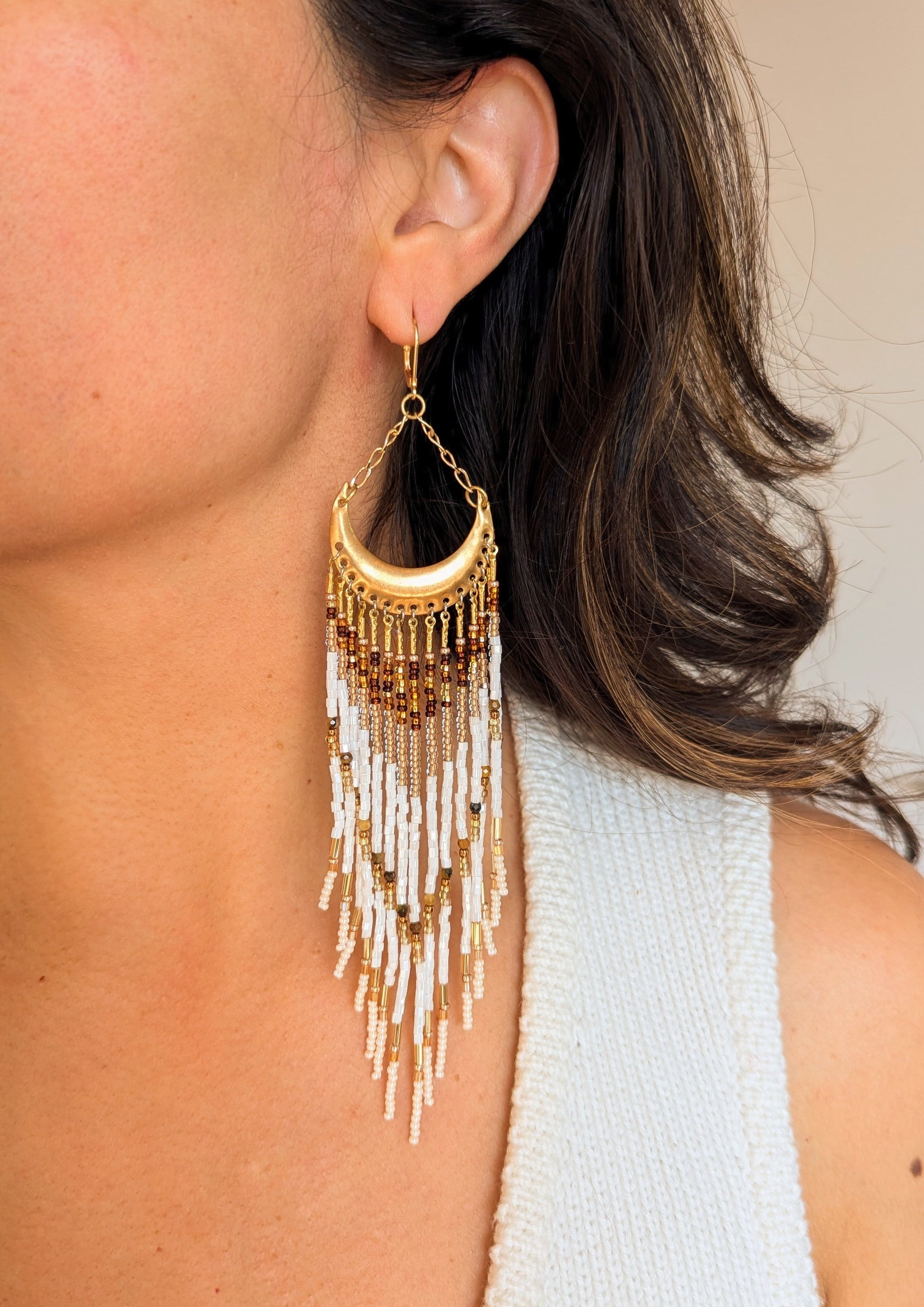 Moon & Milk earring worn on a model, featuring a crescent brass frame with cascading fringes in white, gold, amber, and brown stones.