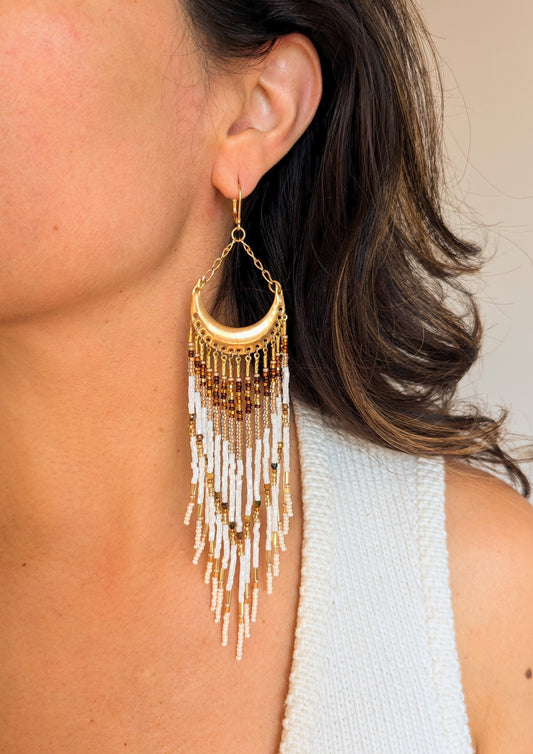 Moon & Milk earring worn on a model, featuring a crescent brass frame with cascading fringes in white, gold, amber, and brown stones.