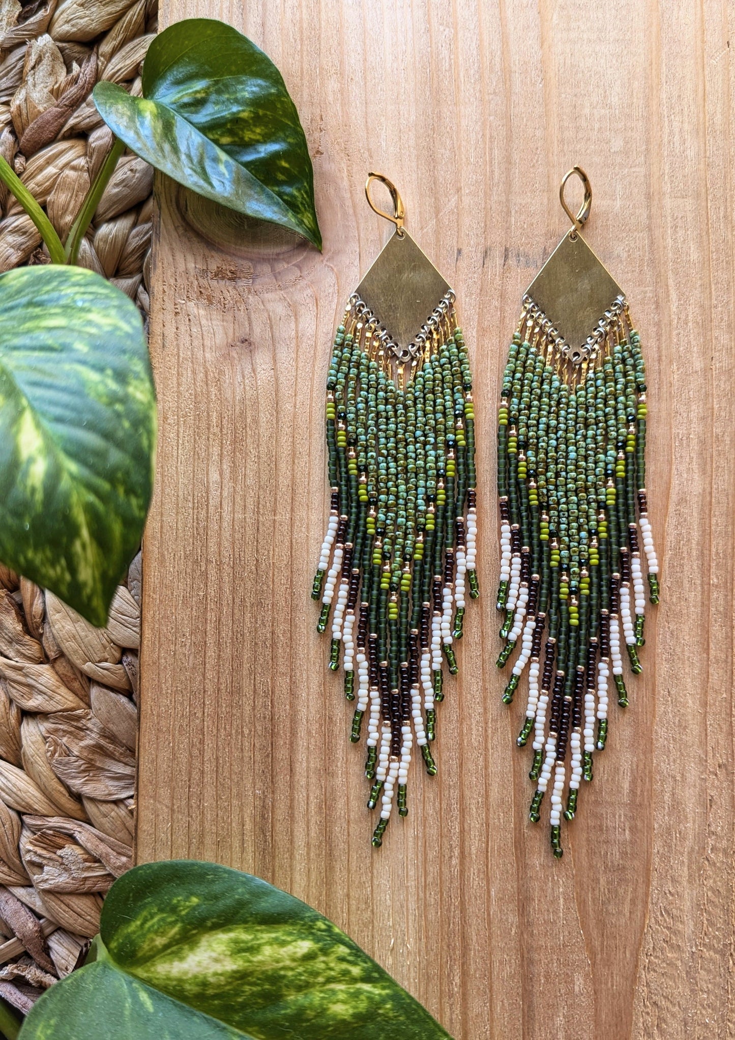 Pair of handmade beaded earrings with brass tops, featuring green, brown, and white beads, laid on a wooden surface next to vibrant green leaves. - Moon & Milk