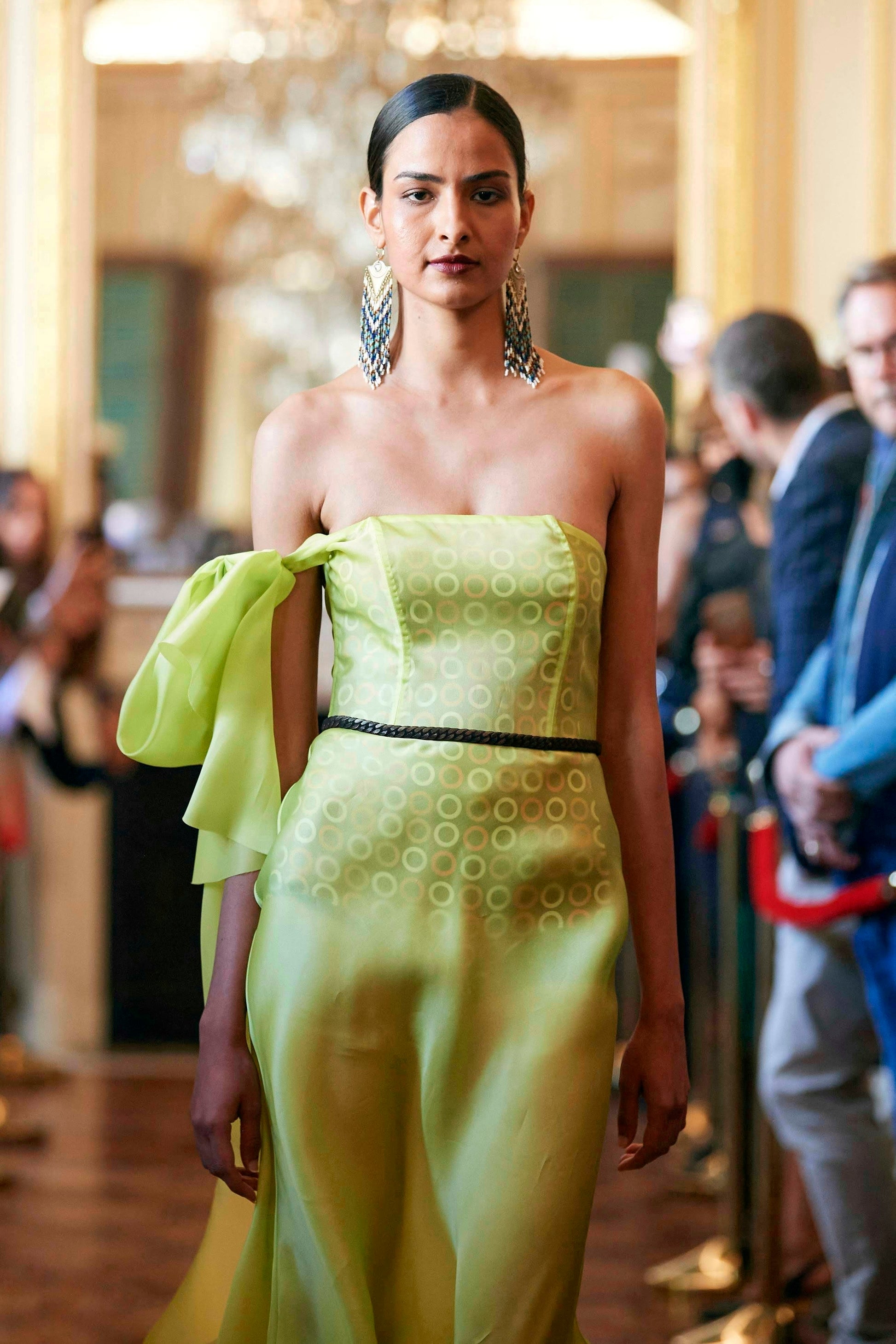 A model wearing bold, handmade beaded fringe earrings with intricate patterns, paired with a strapless lime green dress, walking in a brightly lit venue.