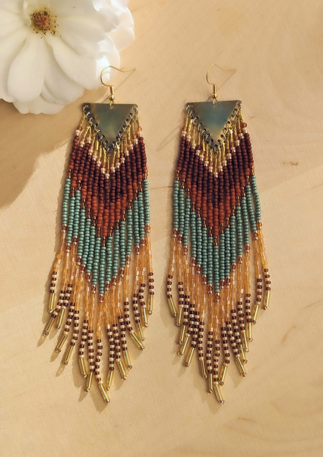 Pair of beaded earrings with a triangular brass top, featuring earthy tones of terracotta, green, and amber with dangling gold tube beads at the ends, placed on a beige background. - Moon & Milk