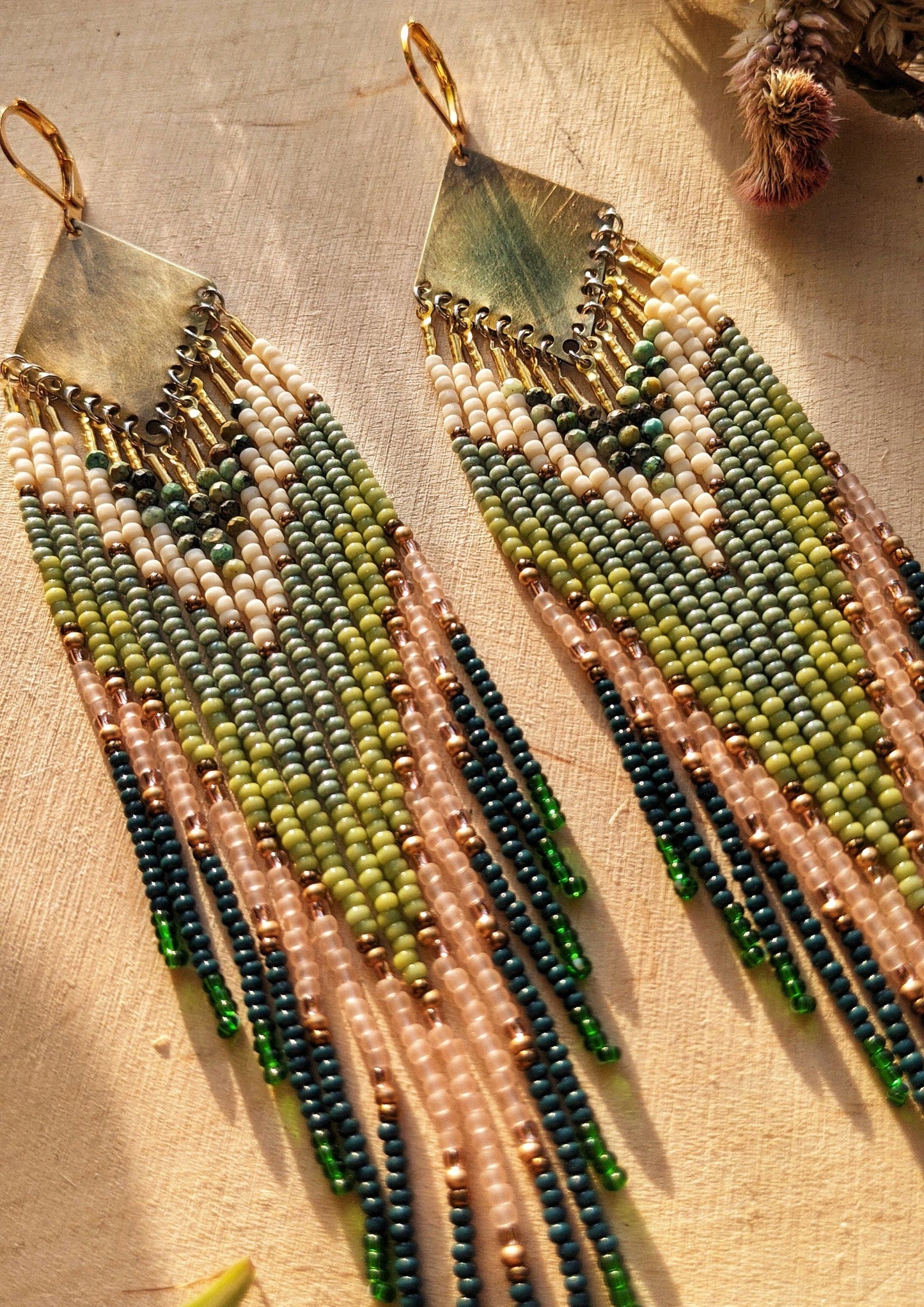 Moon & Milk artisan earrings with layered green, blush, cream, and brass beadwork in a chevron pattern, photographed on textured fabric in warm light.