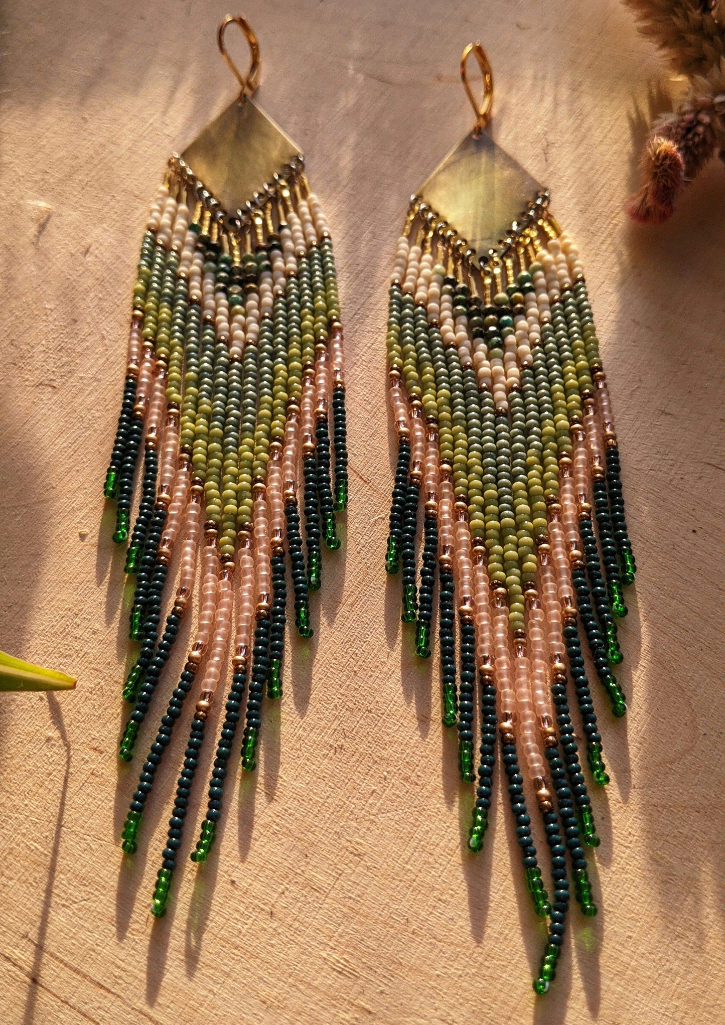 Moon & Milk artisan earrings with layered green, blush, cream, and brass beadwork in a chevron pattern, photographed on textured fabric in warm light.