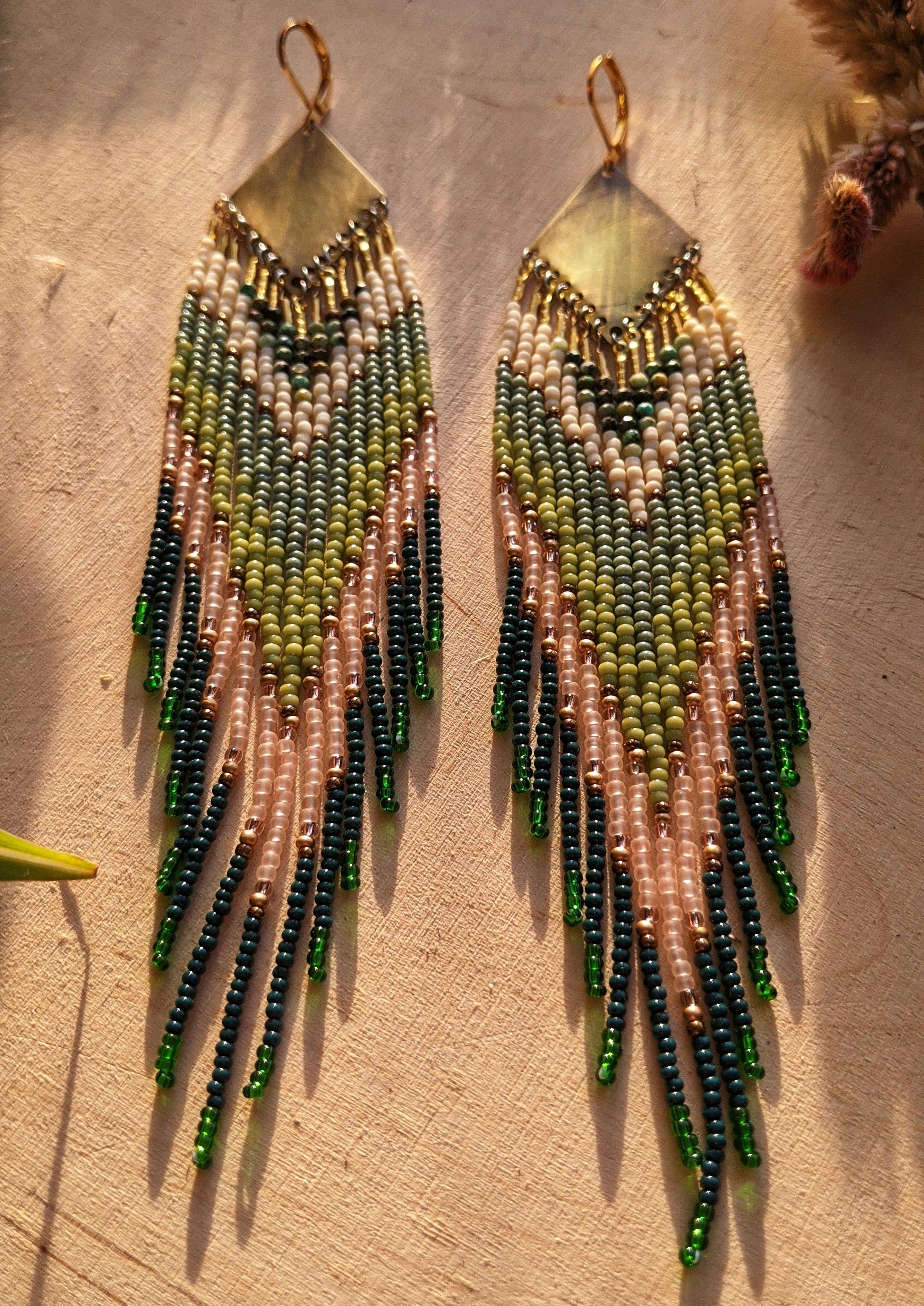 Moon & Milk artisan earrings with layered green, blush, cream, and brass beadwork in a chevron pattern, photographed on textured fabric in warm light.