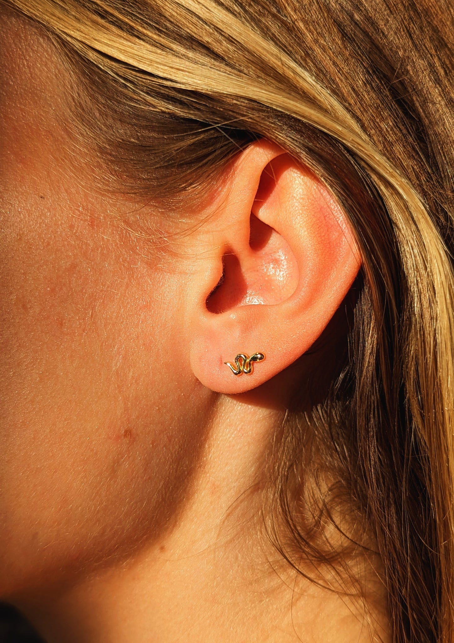 Close-up of a gold snake stud earring by Moon & Milk worn on the earlobe, catching warm sunlight against bare skin and blonde hair.