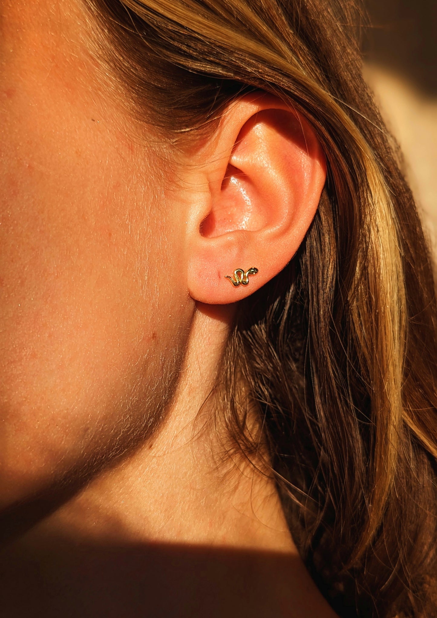 Close-up of a gold snake stud earring by Moon & Milk worn on the earlobe, catching warm sunlight against bare skin and blonde hair.