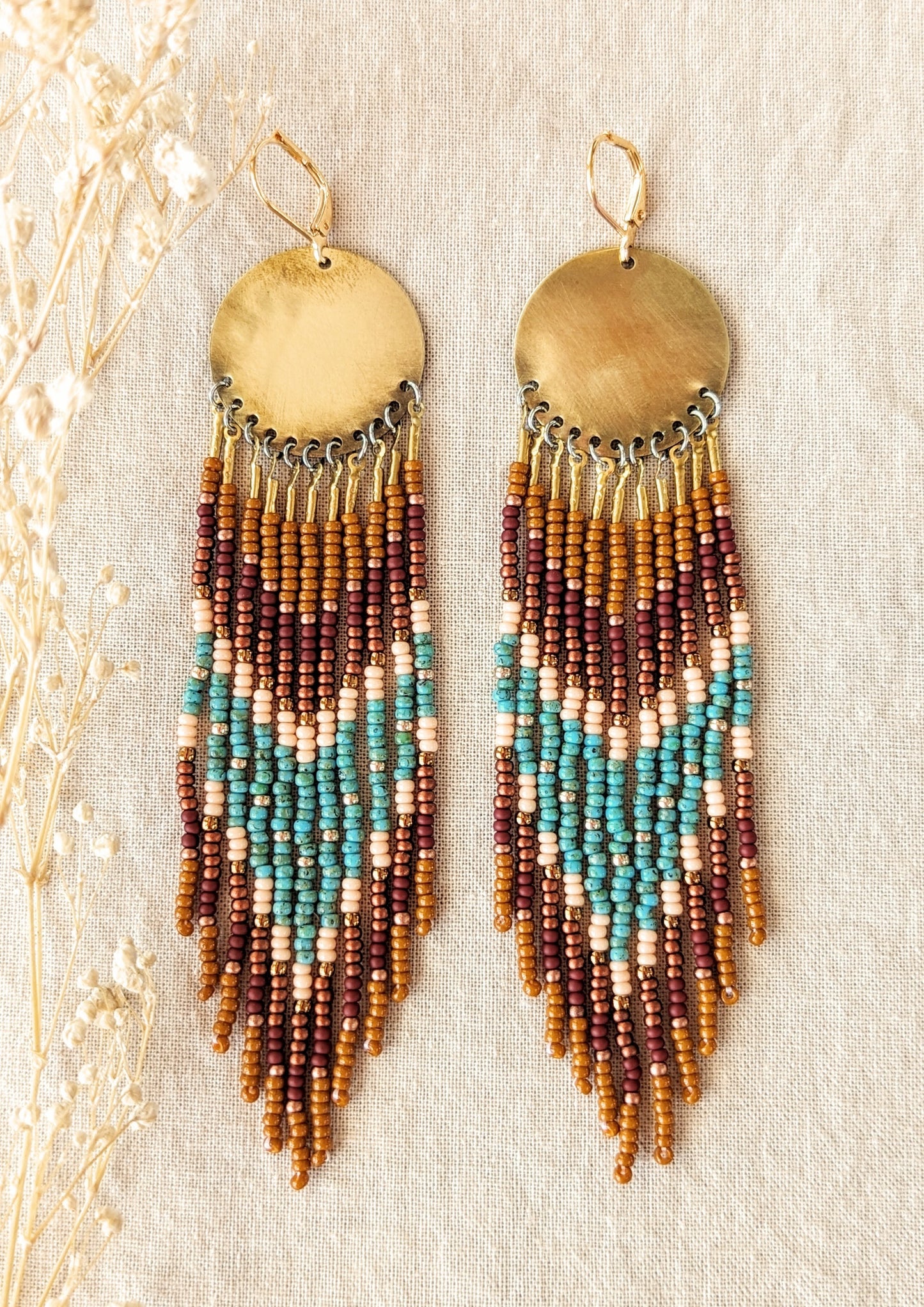 Moon & Milk handmade earrings with brown, turquoise, and peach seed beads hanging from round brass discs, laid on a beige linen surface