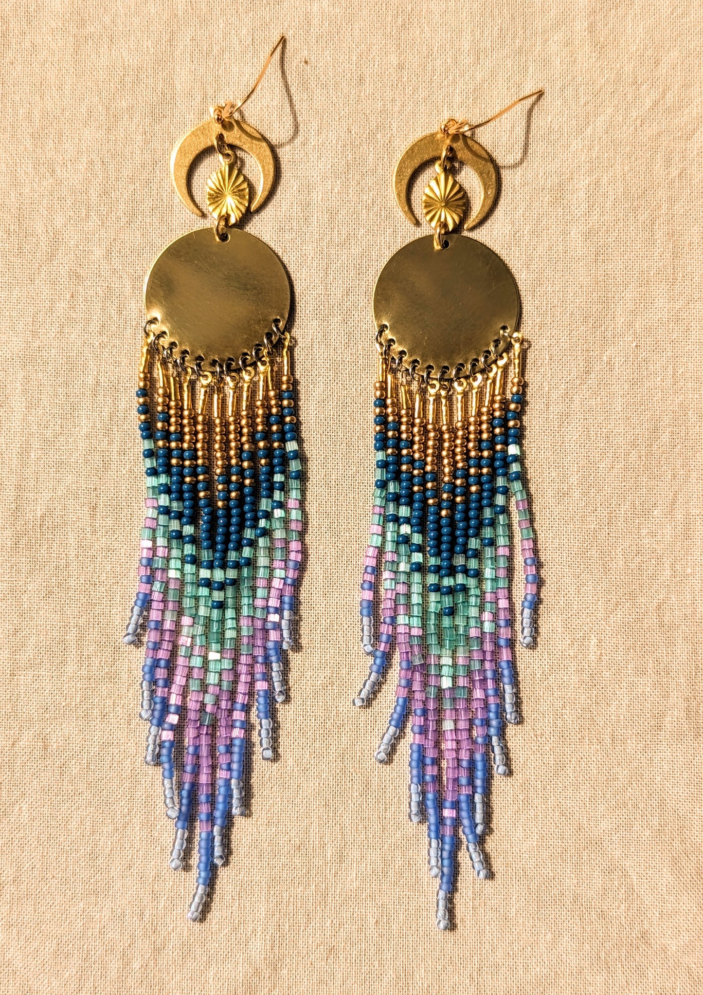 Moon & Milk’s Luna en Tulum earrings with brass moons and gold tops, featuring cascading fringe in navy, lilac, mint, and light blue beads on linen fabric.