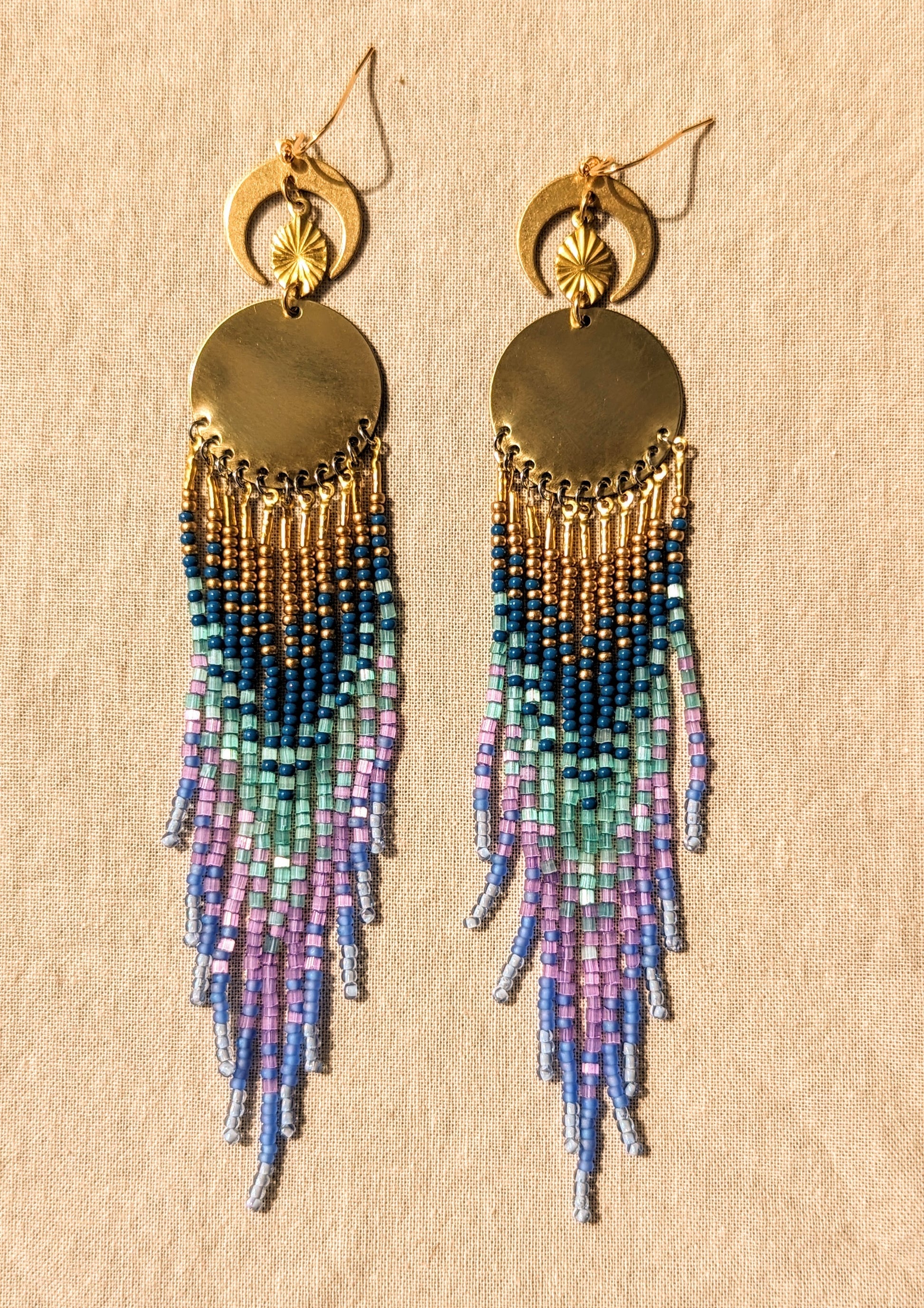 Moon & Milk’s Luna en Tulum earrings with brass moons and gold tops, featuring cascading fringe in navy, lilac, mint, and light blue beads on linen fabric.