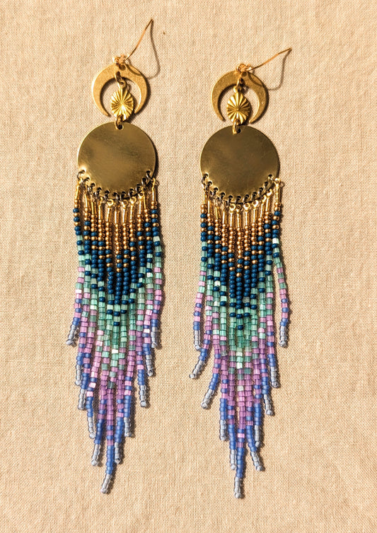 Moon & Milk’s Luna en Tulum earrings with brass moons and gold tops, featuring cascading fringe in navy, lilac, mint, and light blue beads on linen fabric.