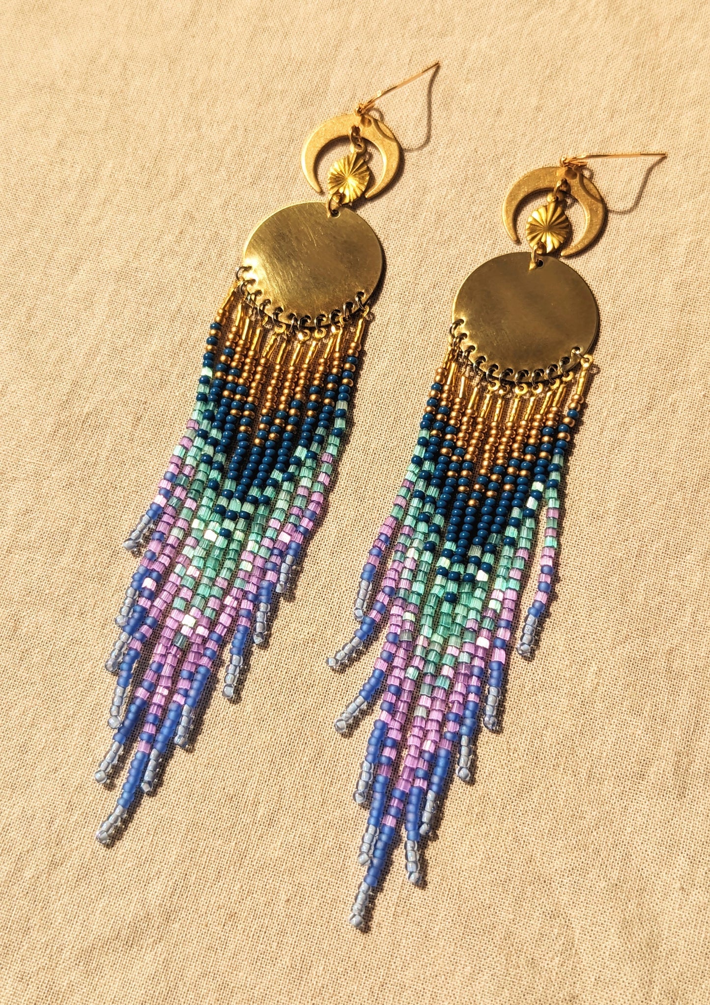 Moon & Milk’s Luna en Tulum earrings with brass moons and gold tops, featuring cascading fringe in navy, lilac, mint, and light blue beads on linen fabric.