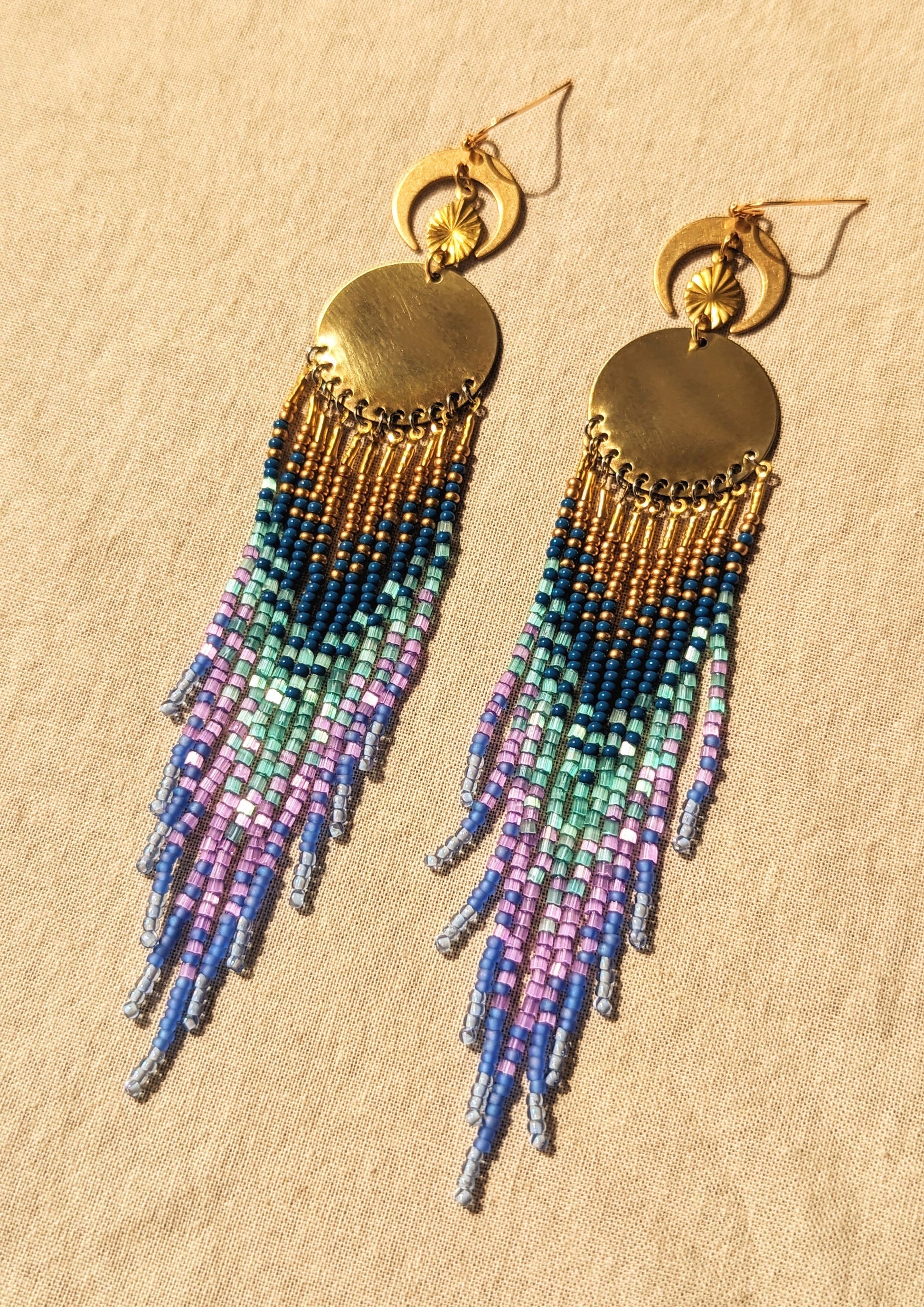 Moon & Milk’s Luna en Tulum earrings with brass moons and gold tops, featuring cascading fringe in navy, lilac, mint, and light blue beads on linen fabric.