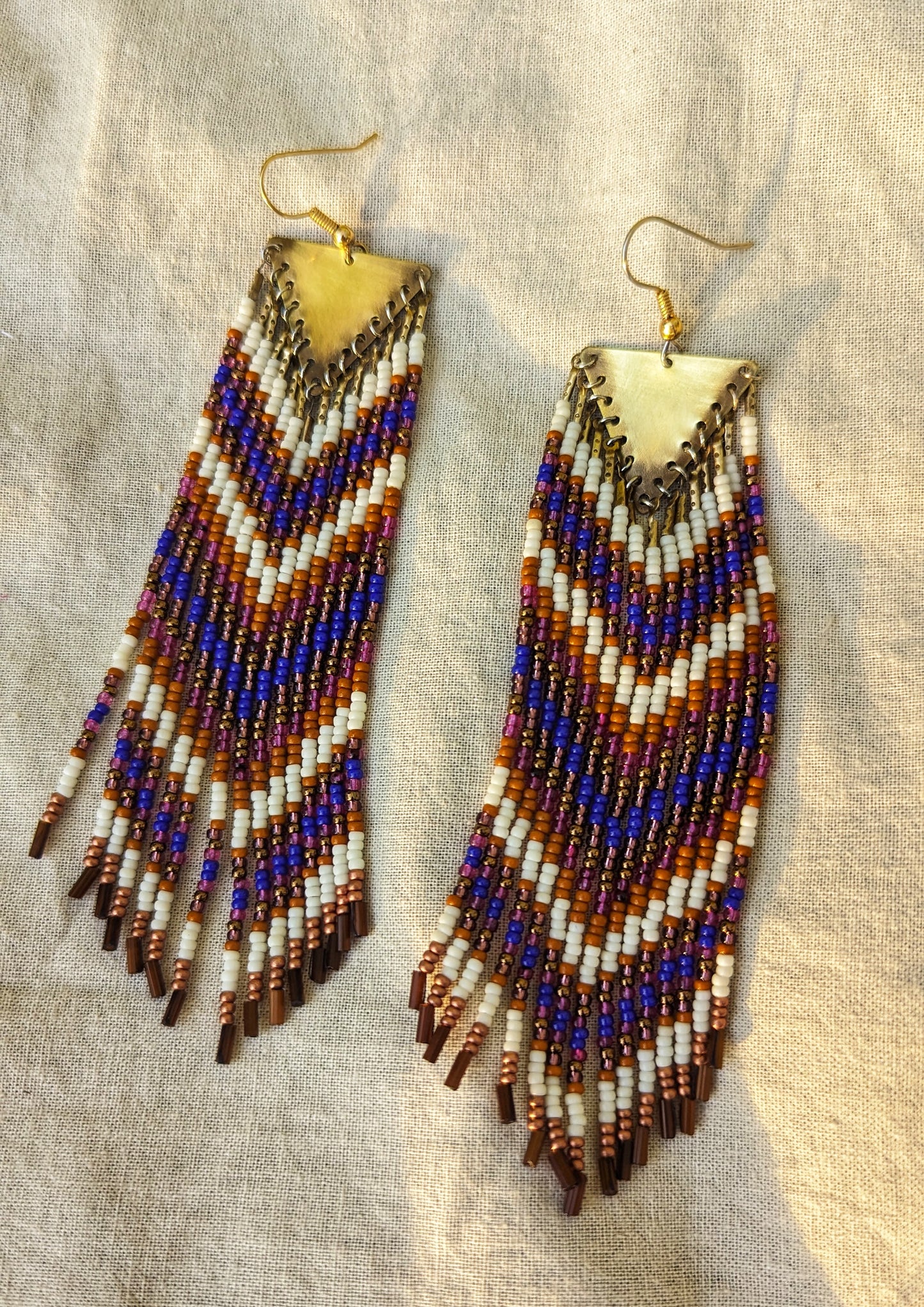 Moon & Milk long native earrings with copper triangle tops and rows of orange, cobalt blue, bronze, and white seed beads on cotton fabric.
