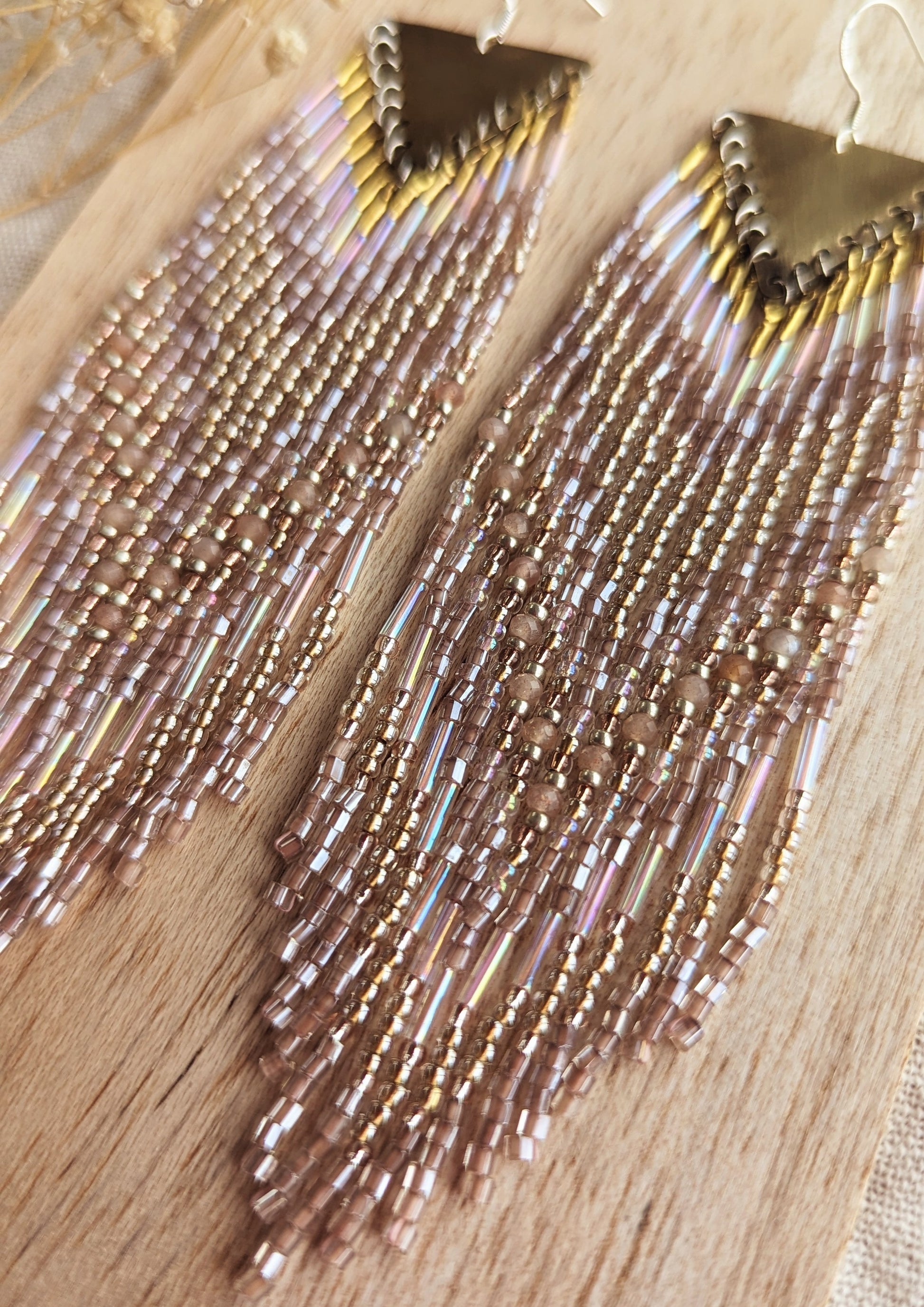 Long native beaded earrings by Moon & Milk featuring brass triangles and soft gold, blush, and iridescent glass beads, laid flat on textured fabric.
