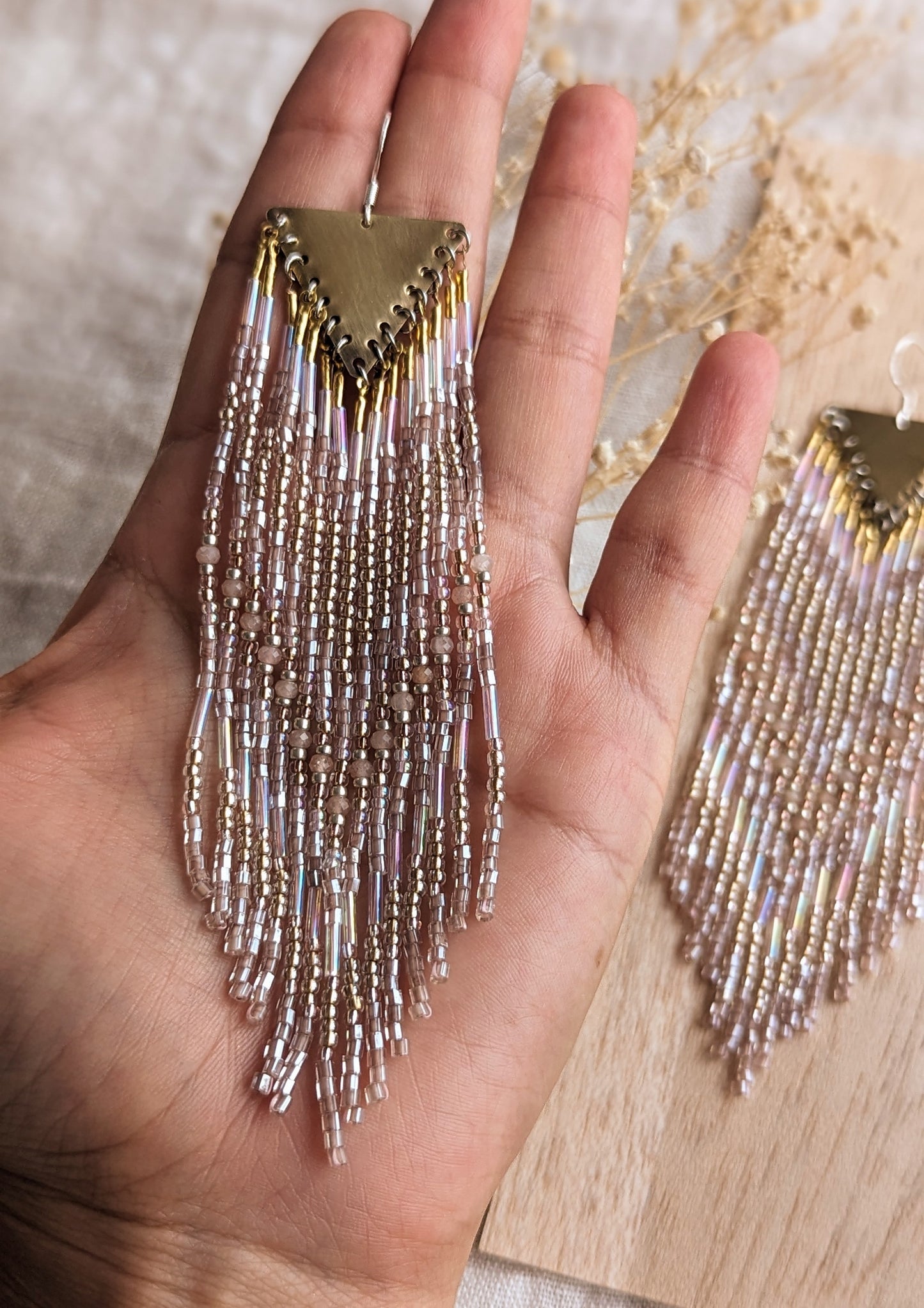Long native beaded earrings by Moon & Milk featuring brass triangles and soft gold, blush, and iridescent glass beads, laid flat on textured fabric.