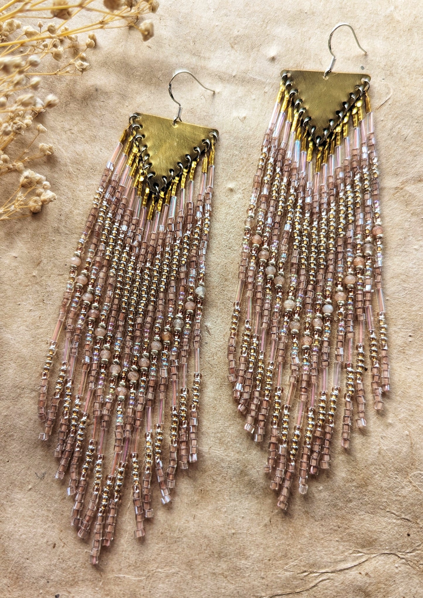 Long native beaded earrings by Moon & Milk featuring brass triangles and soft gold, blush, and iridescent glass beads, laid flat on textured fabric.