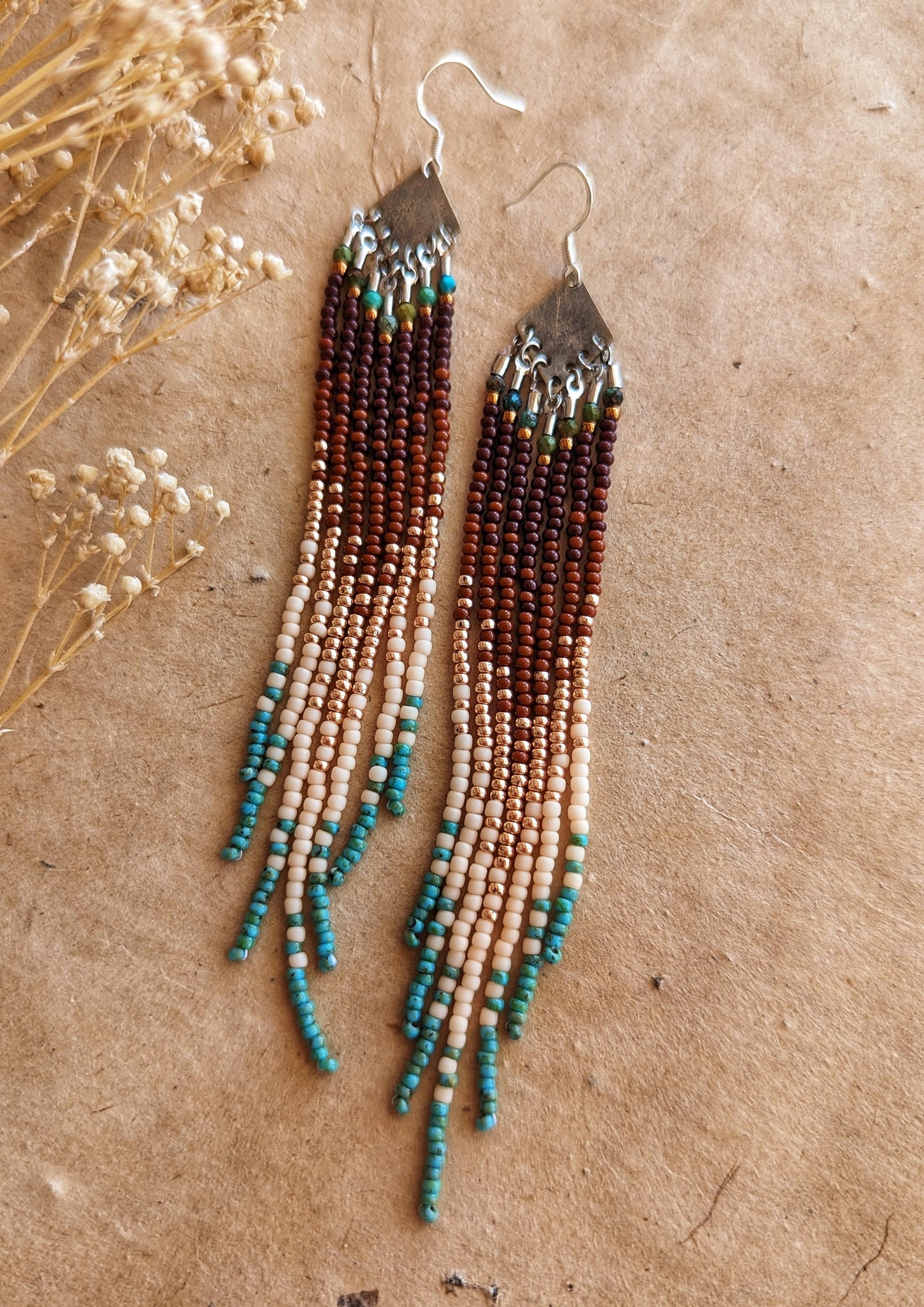 Handcrafted beaded earrings with a cascading fringe design. The beads transition from deep brown and gold to soft beige and turquoise, creating a warm, earthy palette.