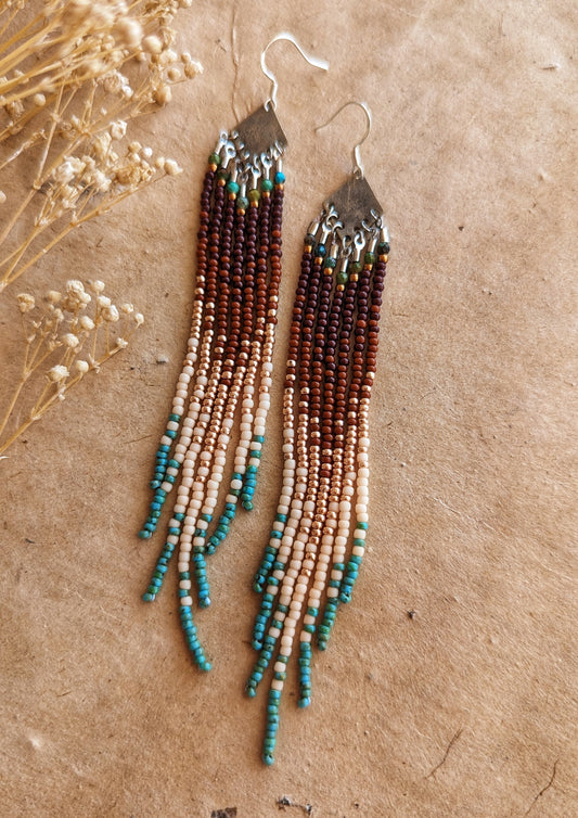 Handcrafted beaded earrings with a cascading fringe design. The beads transition from deep brown and gold to soft beige and turquoise, creating a warm, earthy palette.