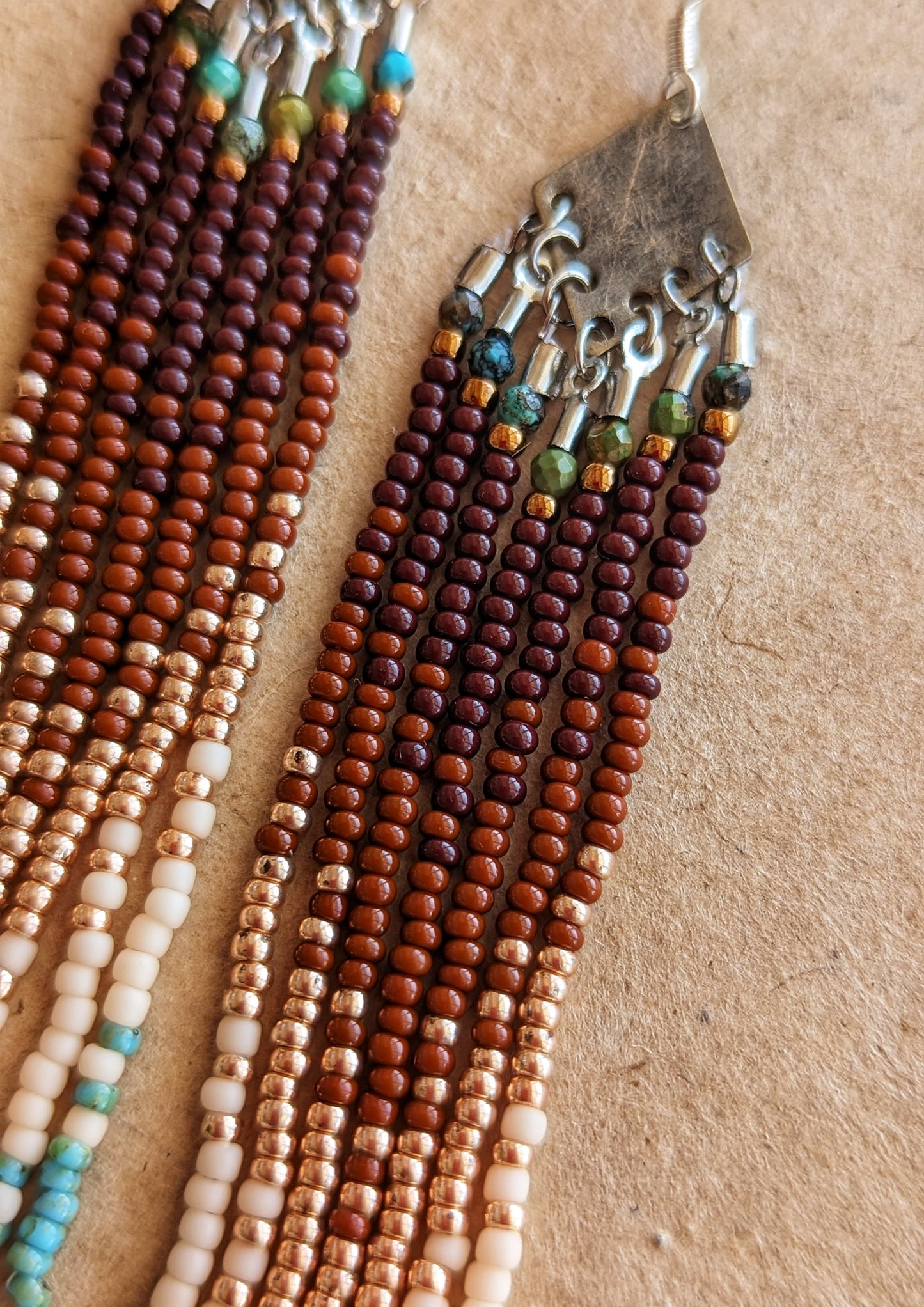 Handcrafted beaded earrings with a cascading fringe design. The beads transition from deep brown and gold to soft beige and turquoise, creating a warm, earthy palette.