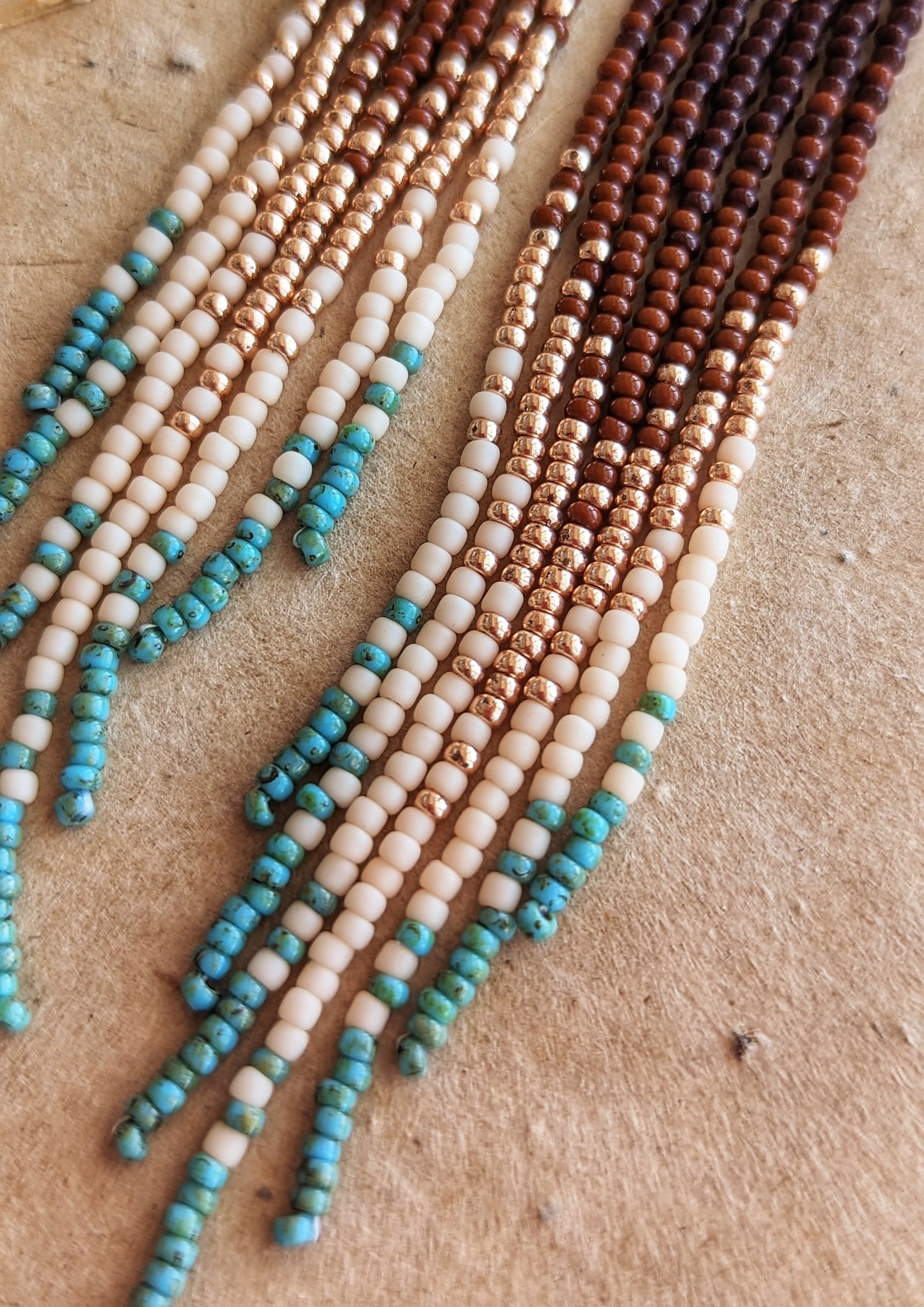 Handcrafted beaded earrings with a cascading fringe design. The beads transition from deep brown and gold to soft beige and turquoise, creating a warm, earthy palette.