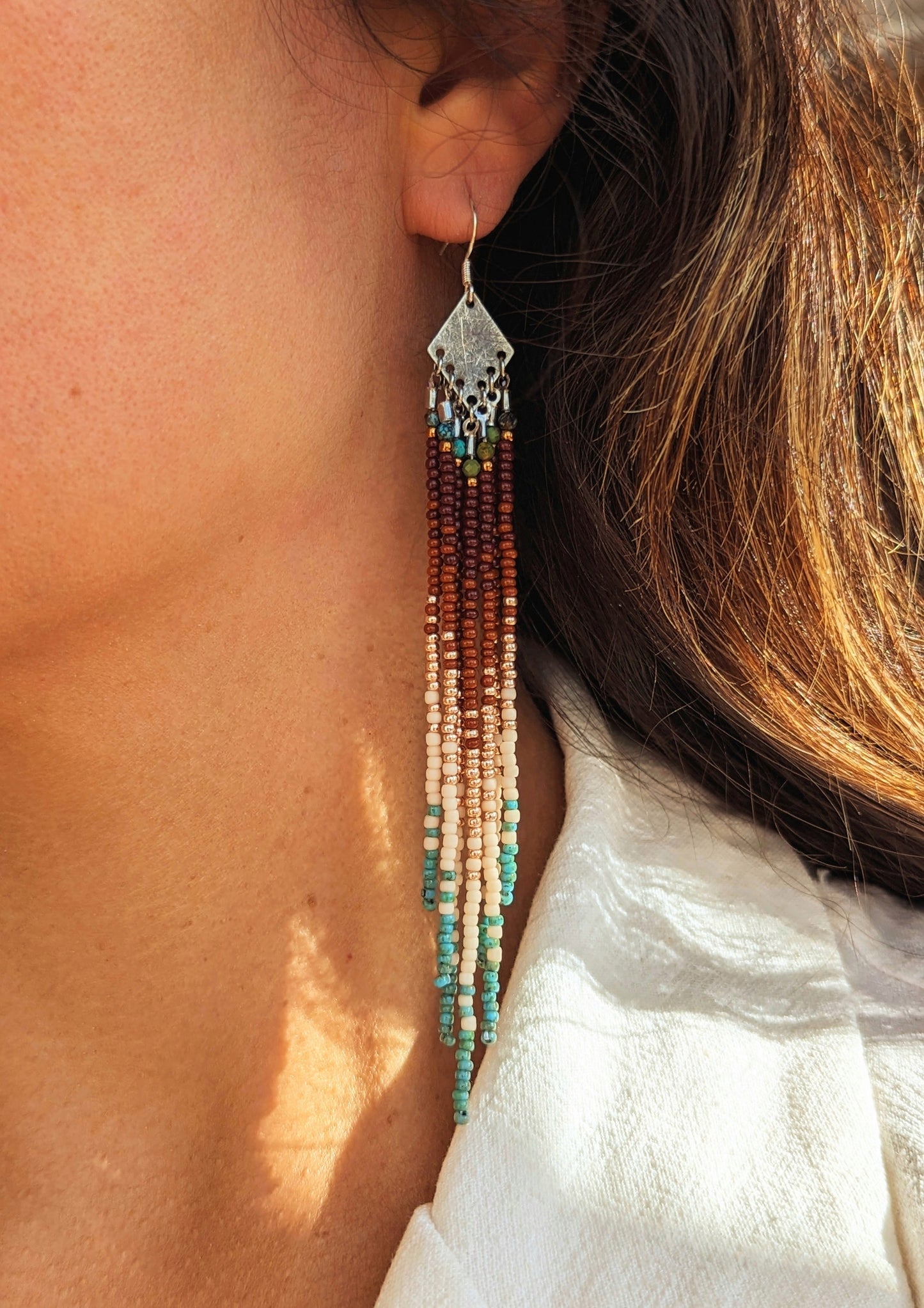 Handcrafted beaded earrings on a model with a cascading fringe design. The beads transition from deep brown and gold to soft beige and turquoise, creating a warm, earthy palette.