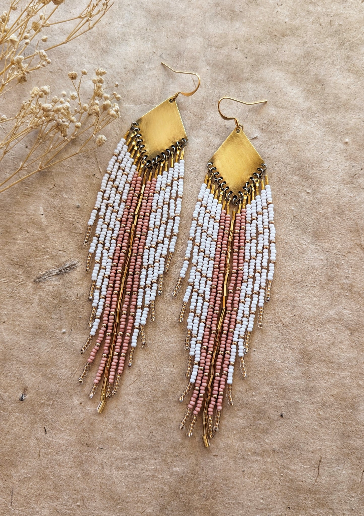 Moon & Milk handmade earrings with soft white, blush, and champagne beads, brass diamond tops, and long beaded fringes among dry flowers.