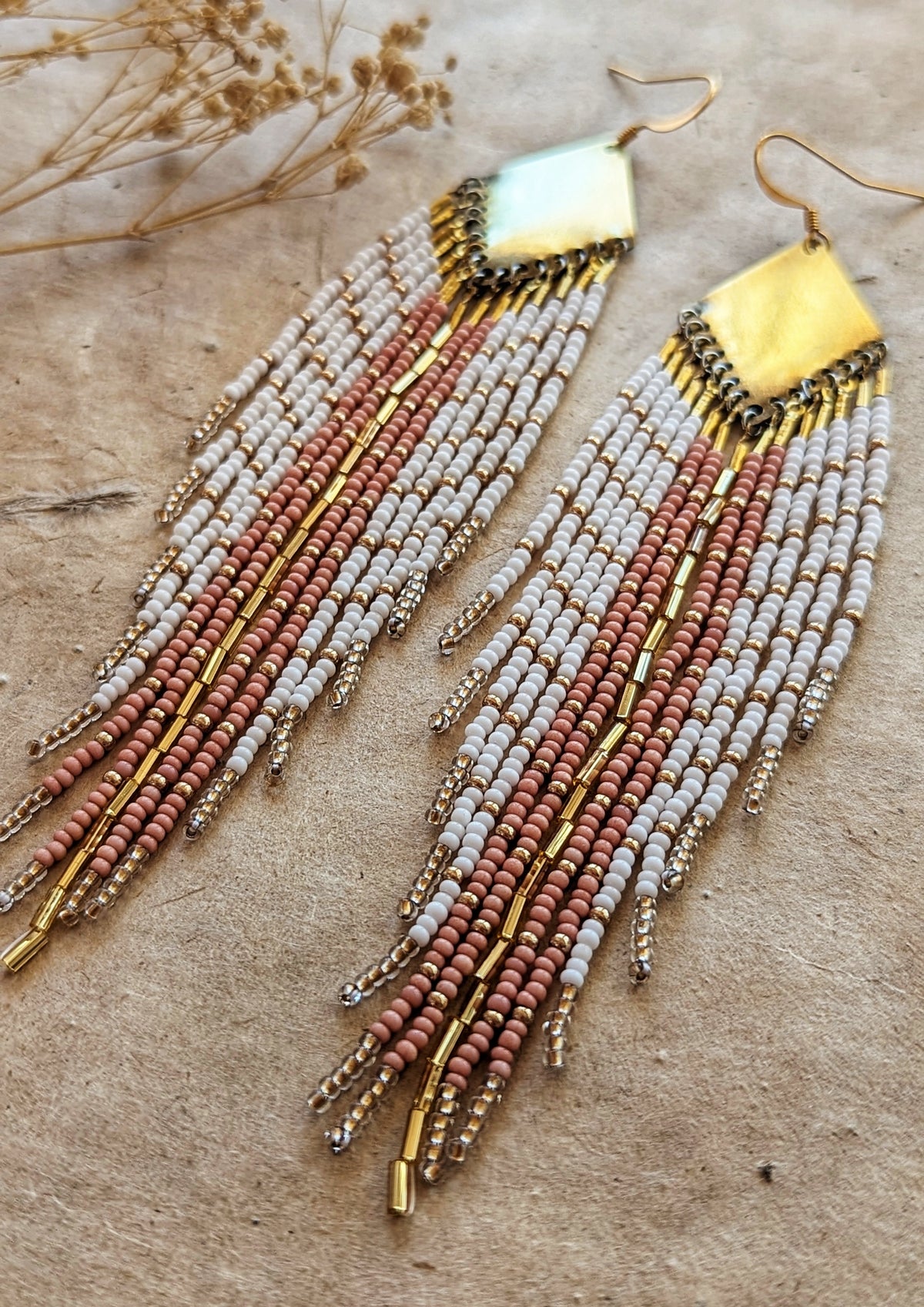 Moon & Milk handmade earrings with soft white, blush, and champagne beads, brass diamond tops, and long beaded fringes among dry flowers.