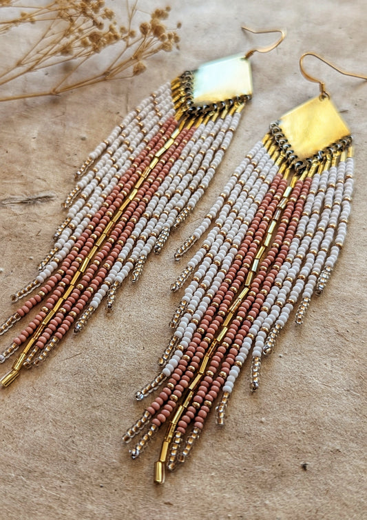 Moon & Milk handmade earrings with soft white, blush, and champagne beads, brass diamond tops, and long beaded fringes among dry flowers.