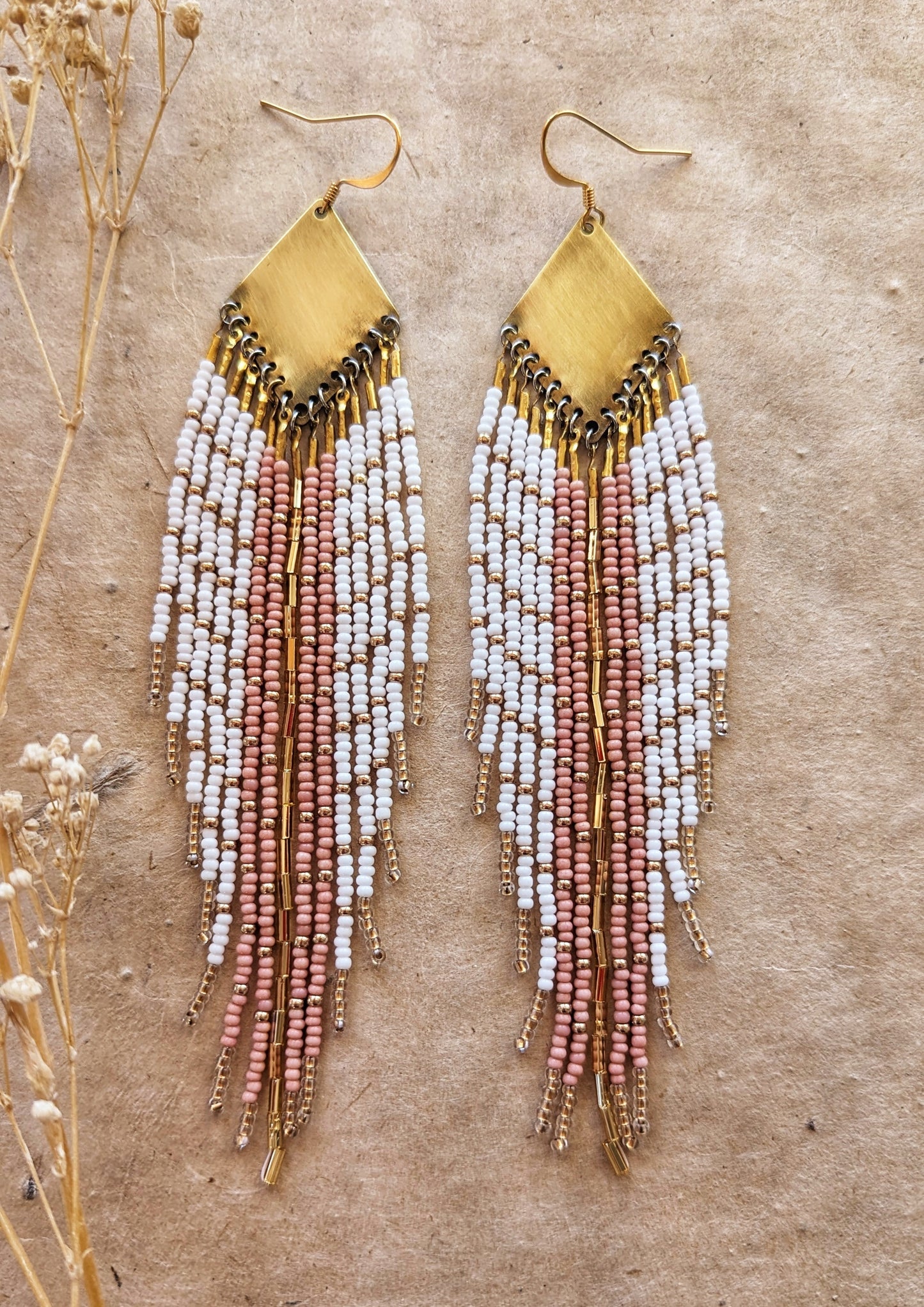 Moon & Milk handmade earrings with soft white, blush, and champagne beads, brass diamond tops, and long beaded fringes among dry flowers