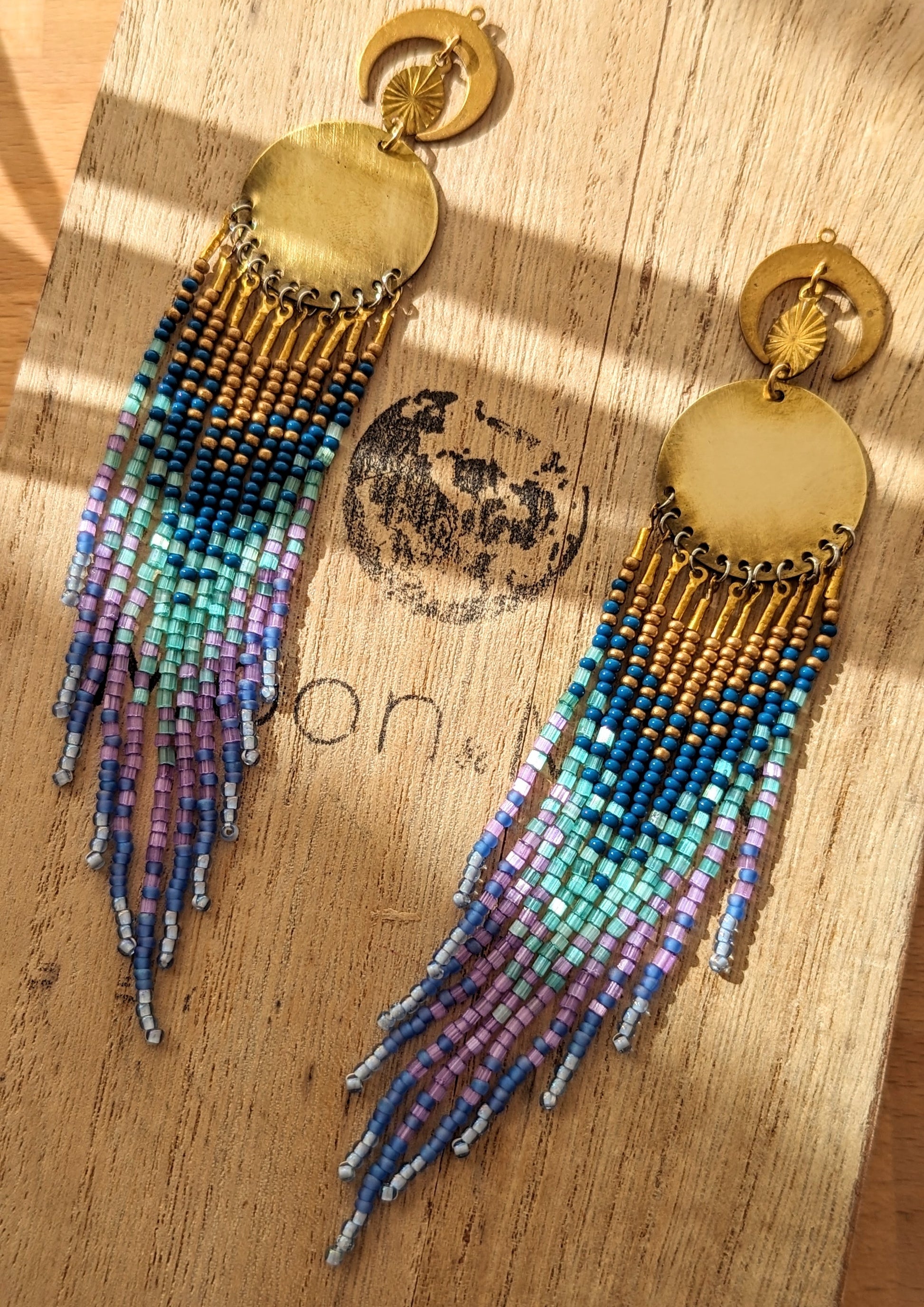 Moon & Milk’s Luna en Tulum earrings with brass moons and gold tops, featuring cascading fringe in navy, lilac, mint, and light blue beads on a wooden box.