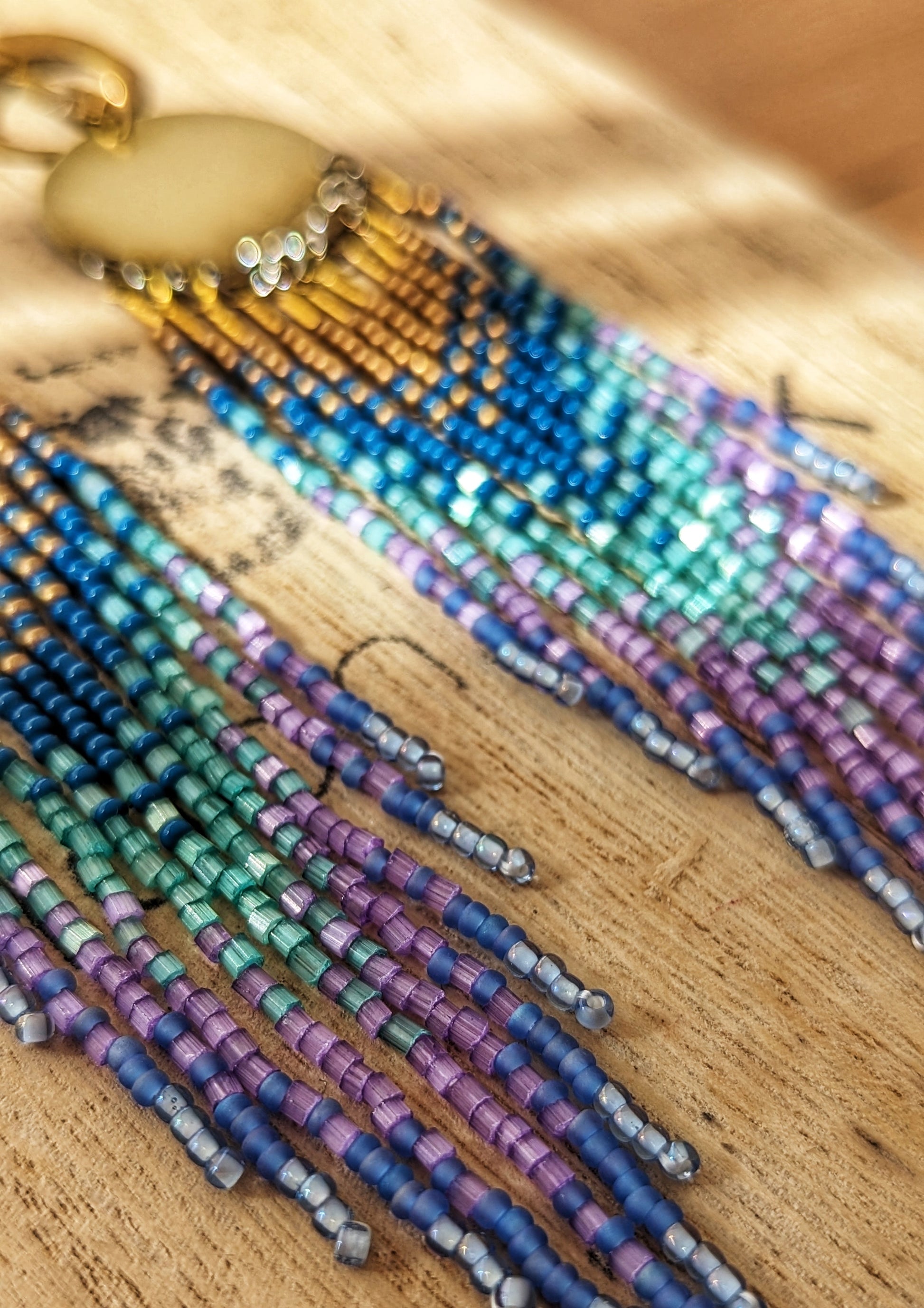 Moon & Milk’s Luna en Tulum earrings with brass moons and gold tops, featuring cascading fringe in navy, lilac, mint, and light blue beads on a wooden box.