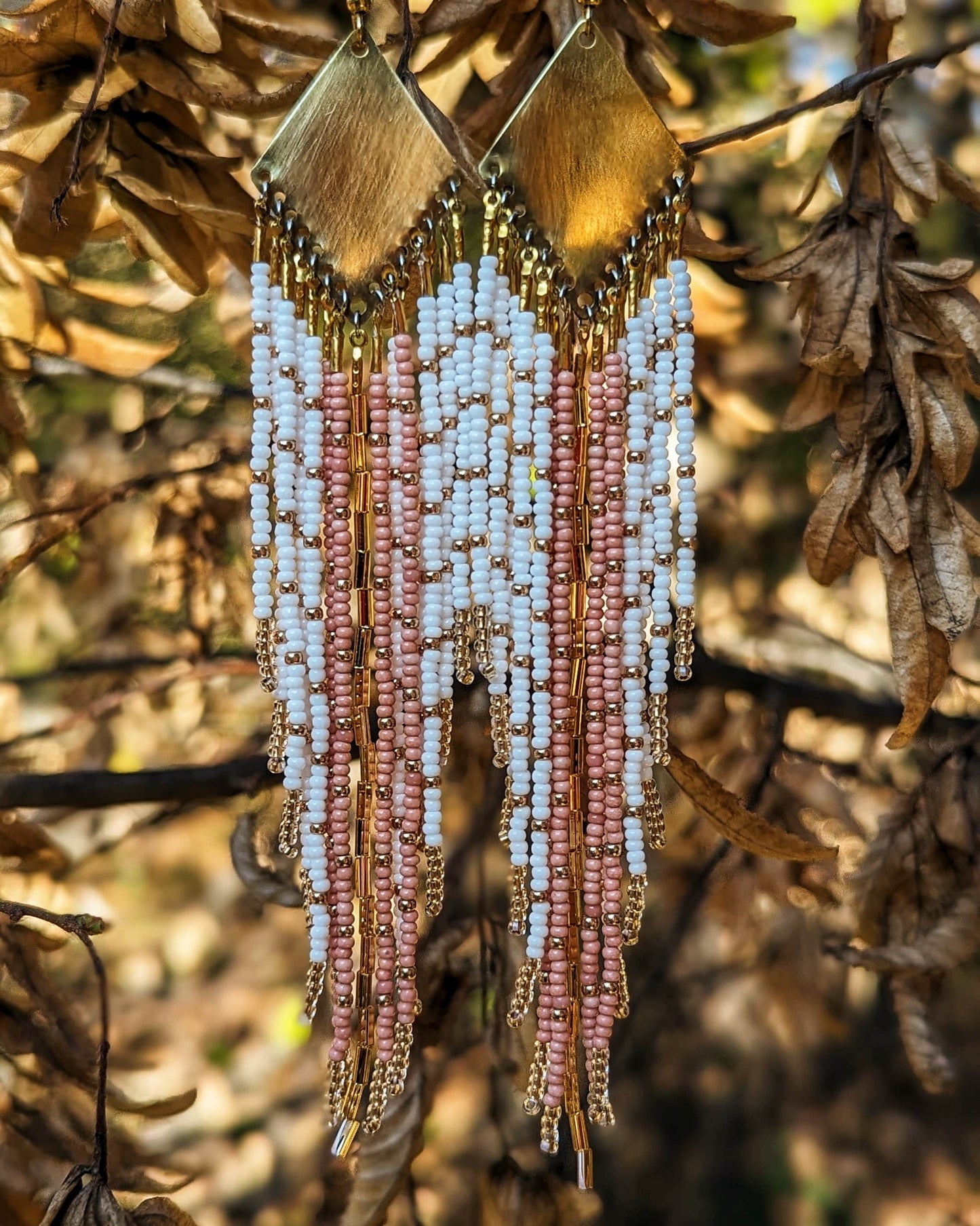 Moon & Milk handmade earrings with soft white, blush, and champagne beads, brass diamond tops, and long beaded fringe hanging among dry leaves.