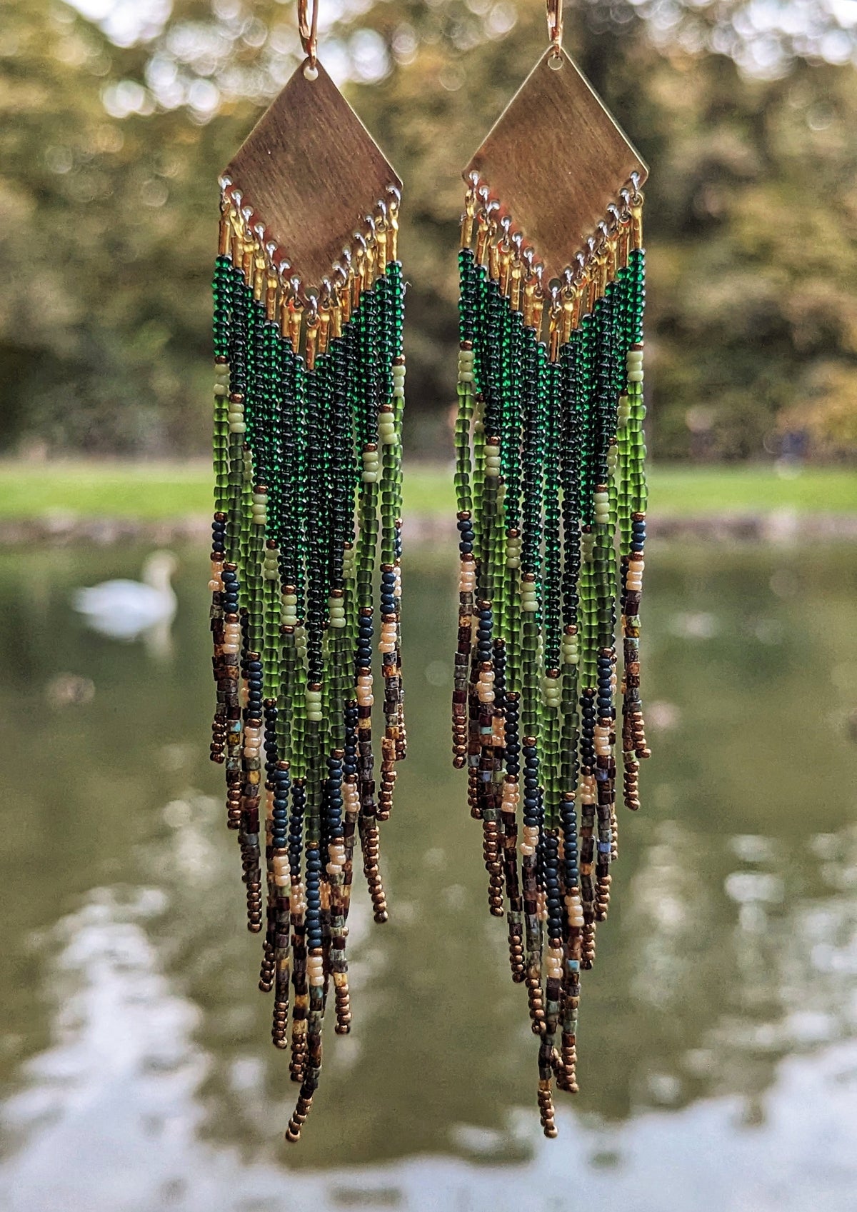 Long native beaded earrings by Moon & Milk in layered green, brown, and matte earthy tones with brass tops, photographed outdoors near a serene pond.
