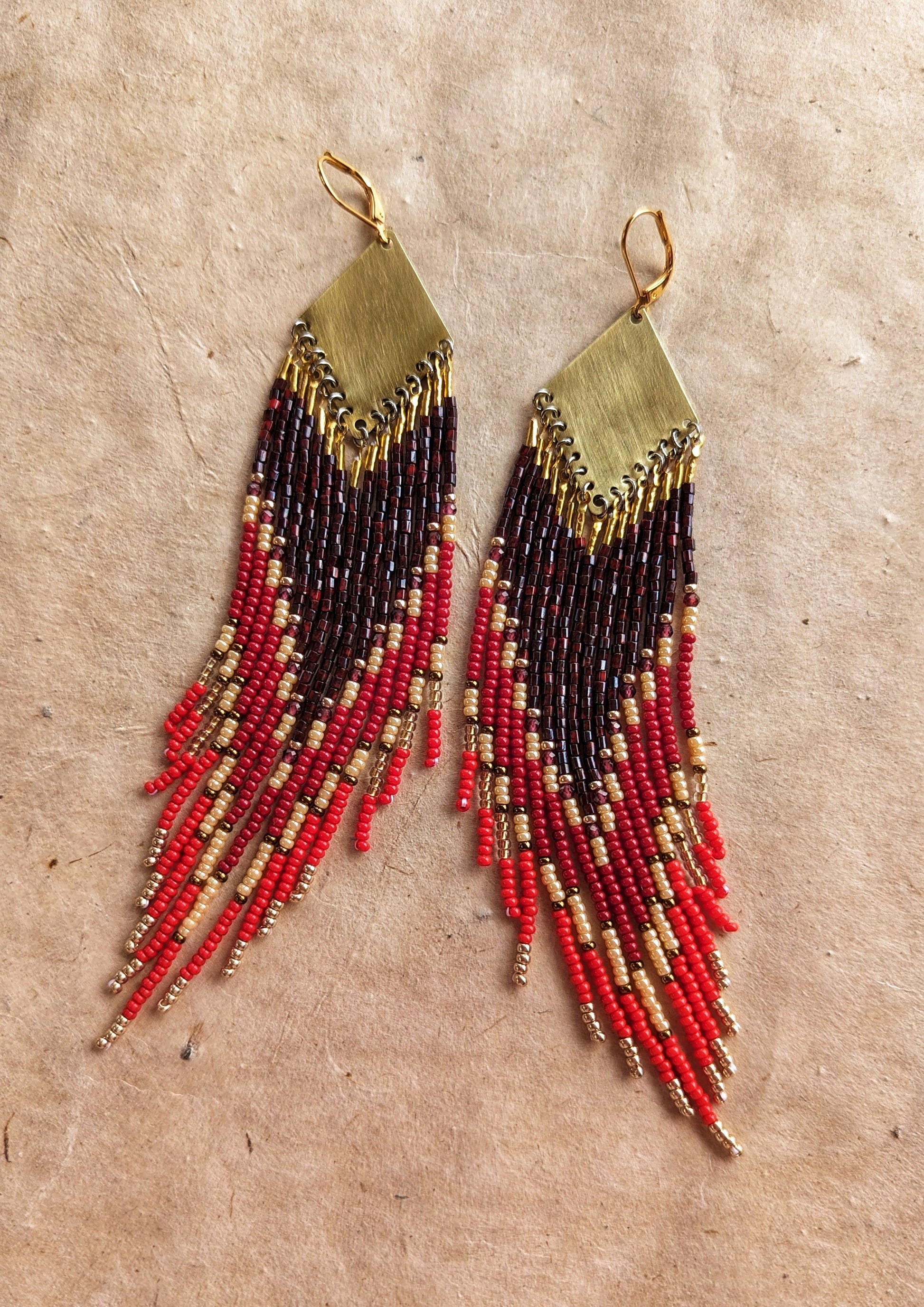 Moon & Milk handmade beaded earrings with crimson, red, and gold beaded fringes cascading from brass diamond tops, resting on sunlit textured parchment.