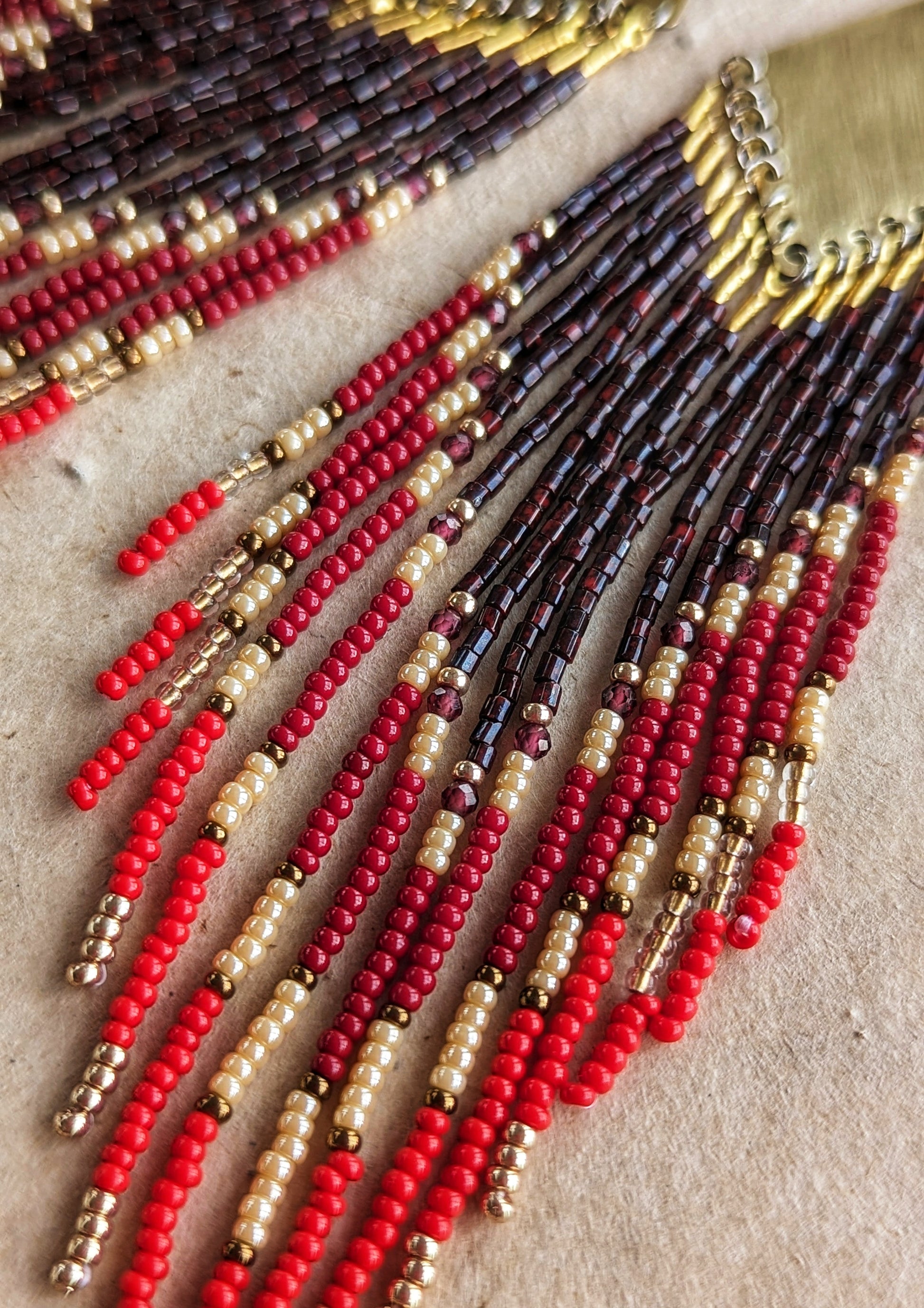 Close-up of handmade beaded earrings with crimson, red, and gold beaded fringes cascading from brass diamond tops, resting on sunlit textured parchment.